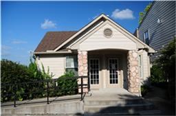 Charming single-story home with a gabled roof, stone accents, and double French doors. Features a front ramp for easy access and is surrounded by lush greenery, offering a cozy and inviting exterior.