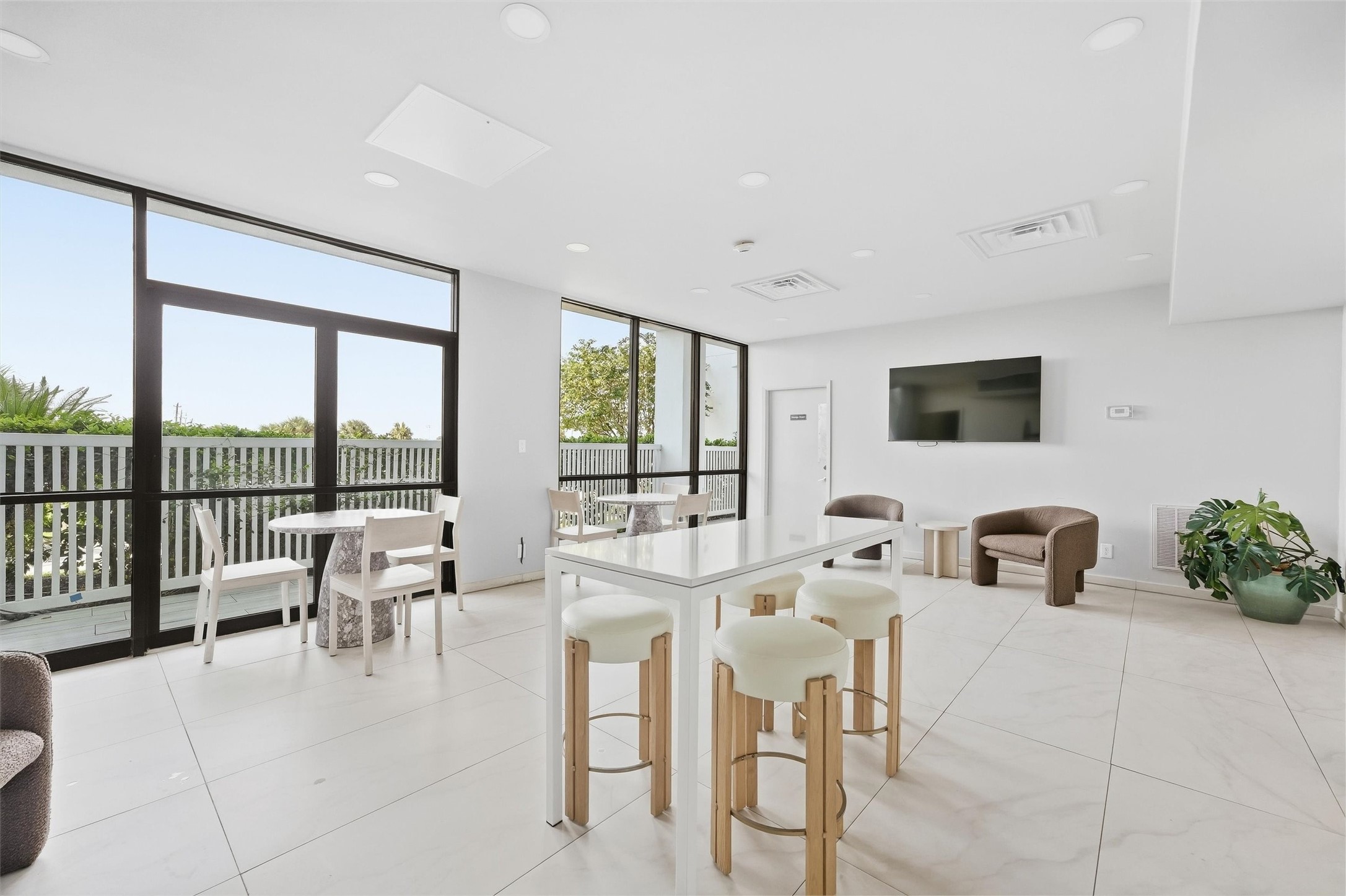Bright and modern living area featuring large windows, sleek tile flooring, and a versatile open layout with a stylish table and comfortable seating, perfect for relaxation or entertaining.