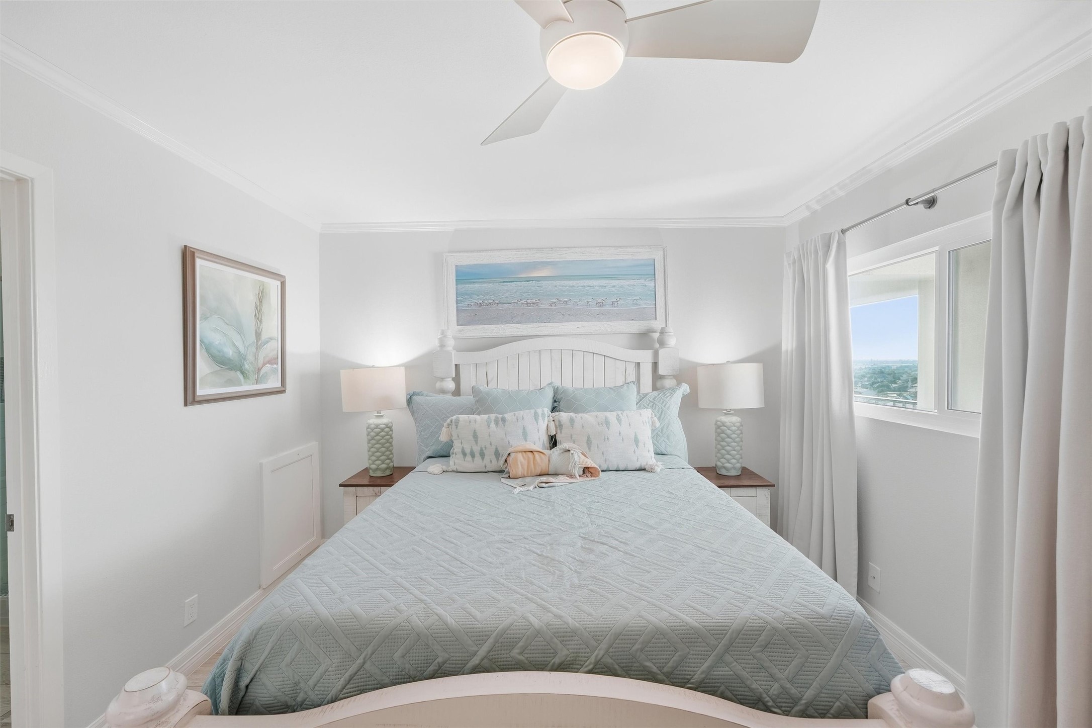 Bright and serene bedroom featuring a comfortable bed with stylish bedding, coastal artwork, and ample natural light from a large window.