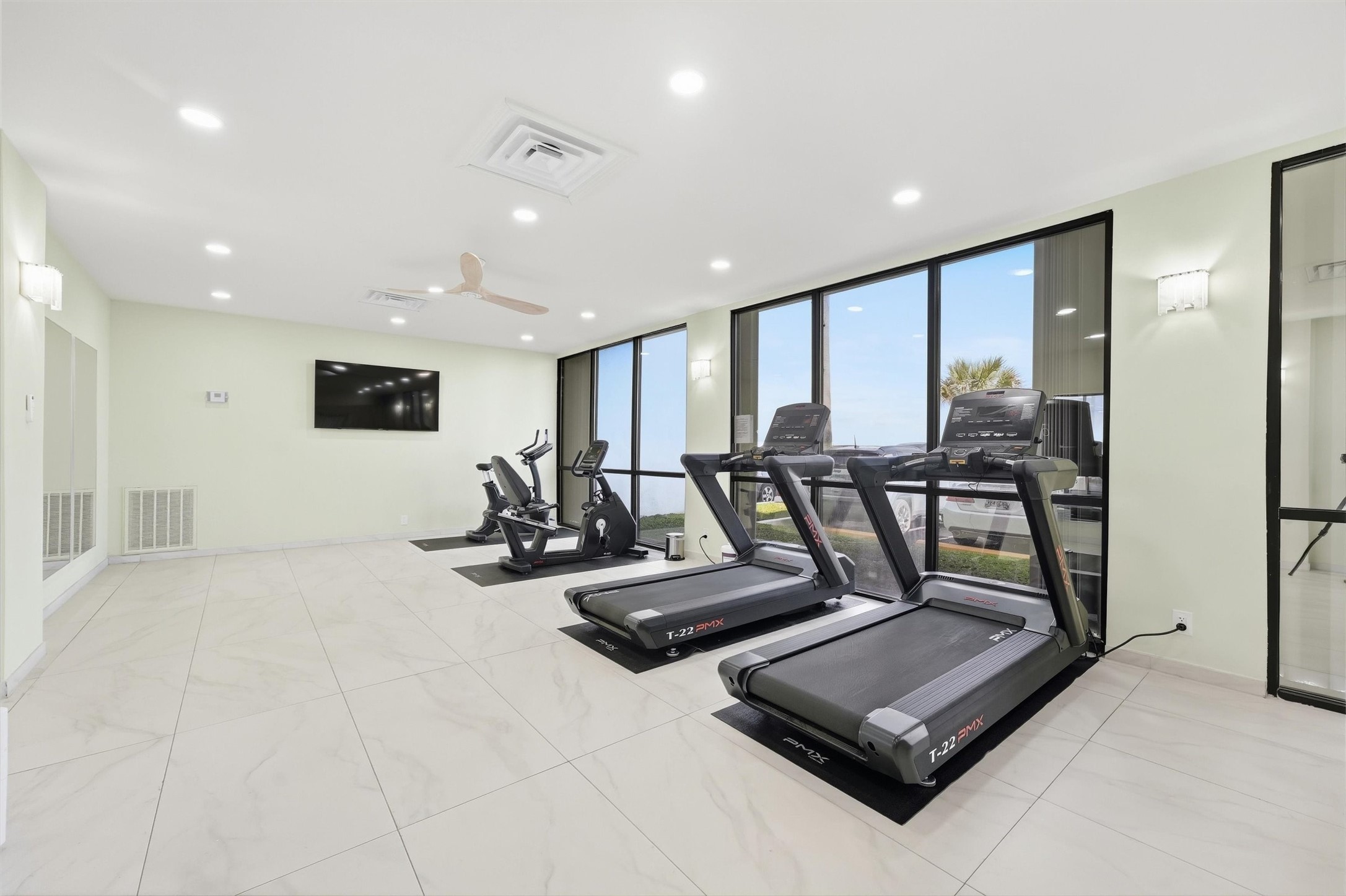 Modern fitness center with large windows, featuring treadmills, exercise bikes, and a wall-mounted TV for an enjoyable workout experience.