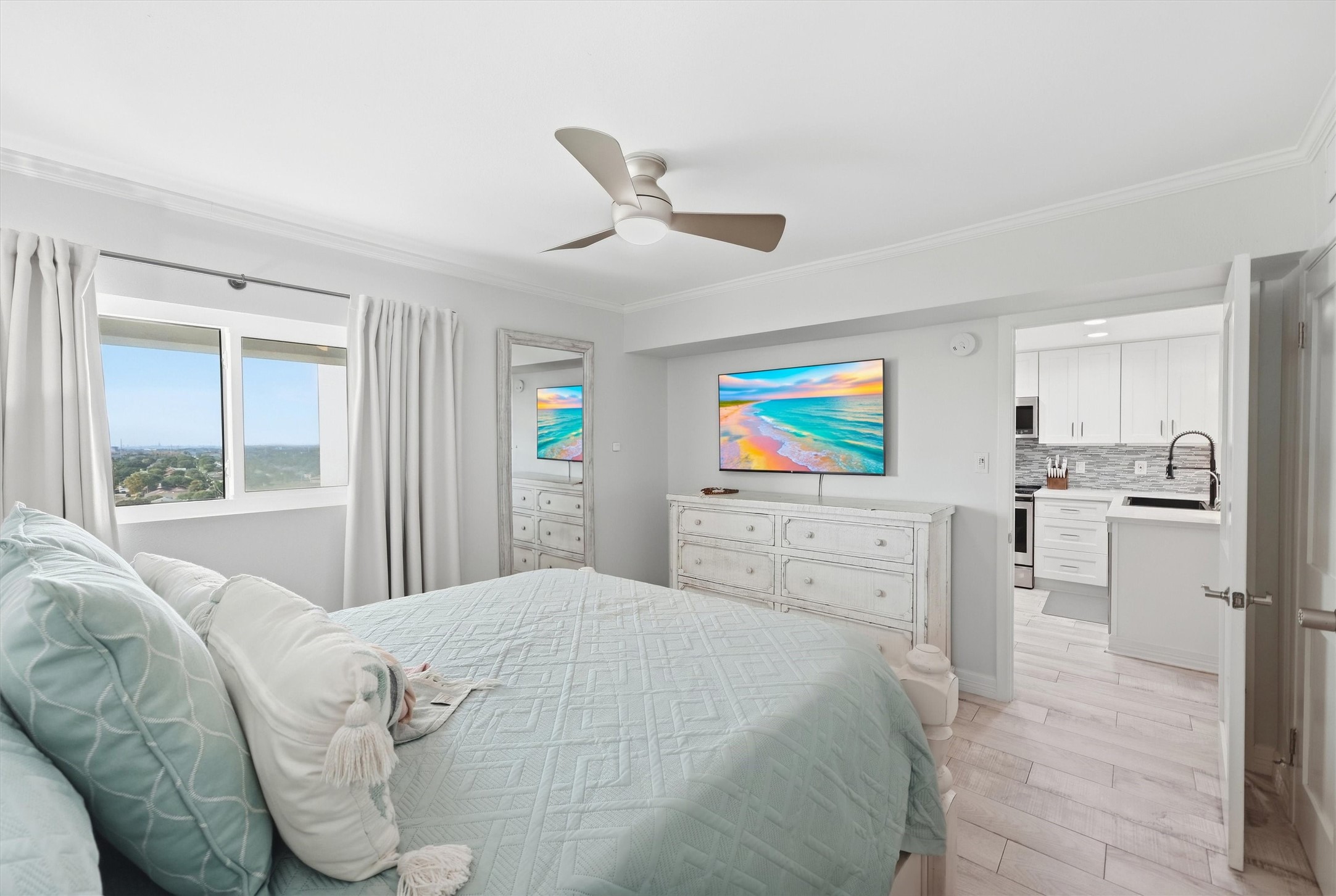 Bright and airy bedroom with large window views, a cozy bed with soft pastel linens, and modern ceiling fan. The room opens into a sleek, white kitchen, adding a sense of spaciousness and flow.