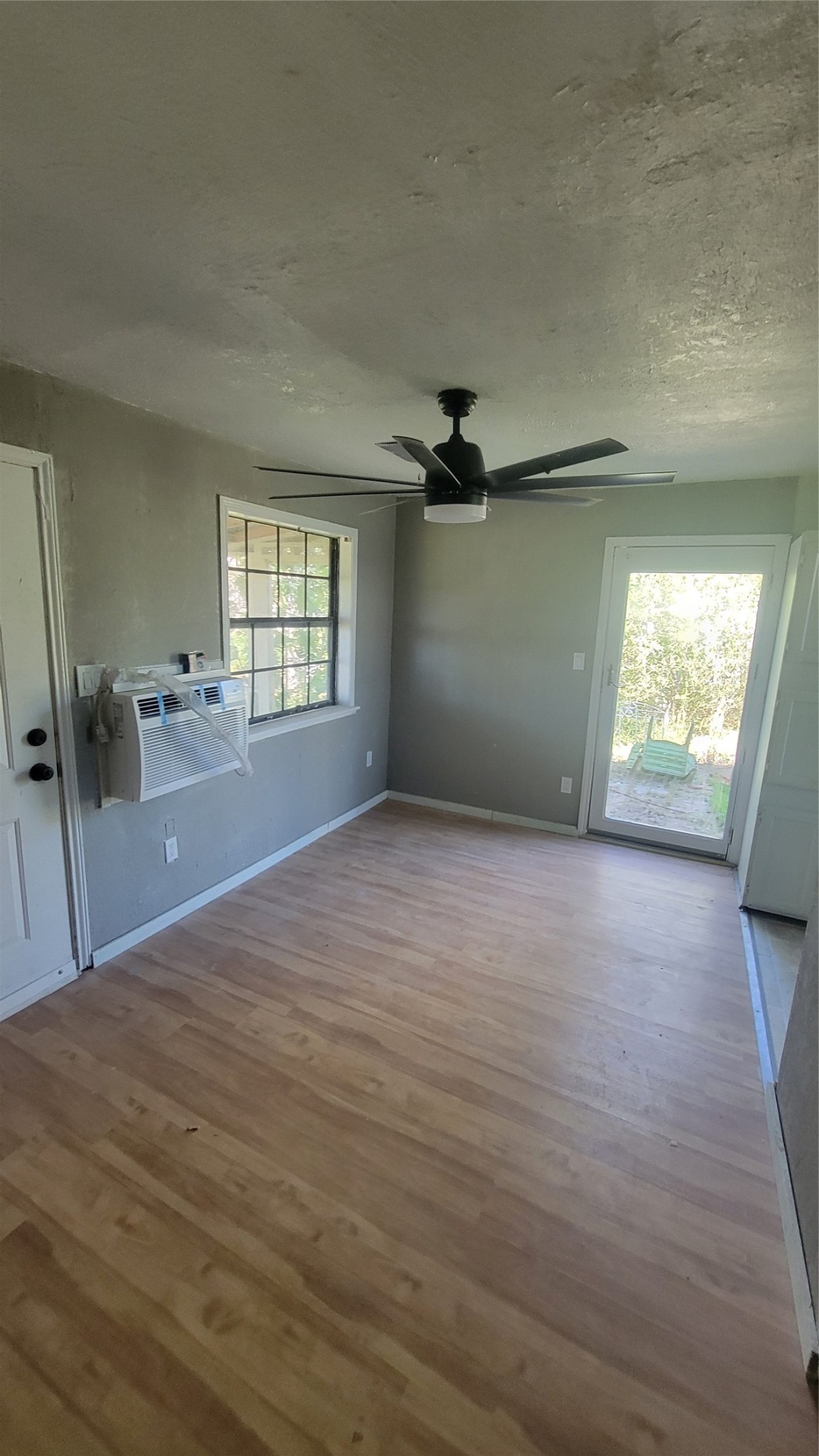 This room features hardwood floors, a ceiling fan, and a wall-mounted air conditioning unit. It has a window for natural light and a door leading to an outdoor area. Ideal for a cozy living space or office.