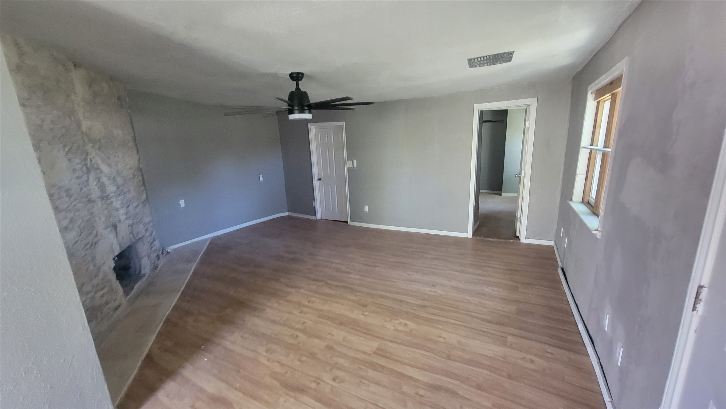 This room features laminate wood flooring, a stone accent wall, and a ceiling fan. It has a neutral color scheme and ample natural light from the window. Theres a doorway leading to another room, adding to the open feel.