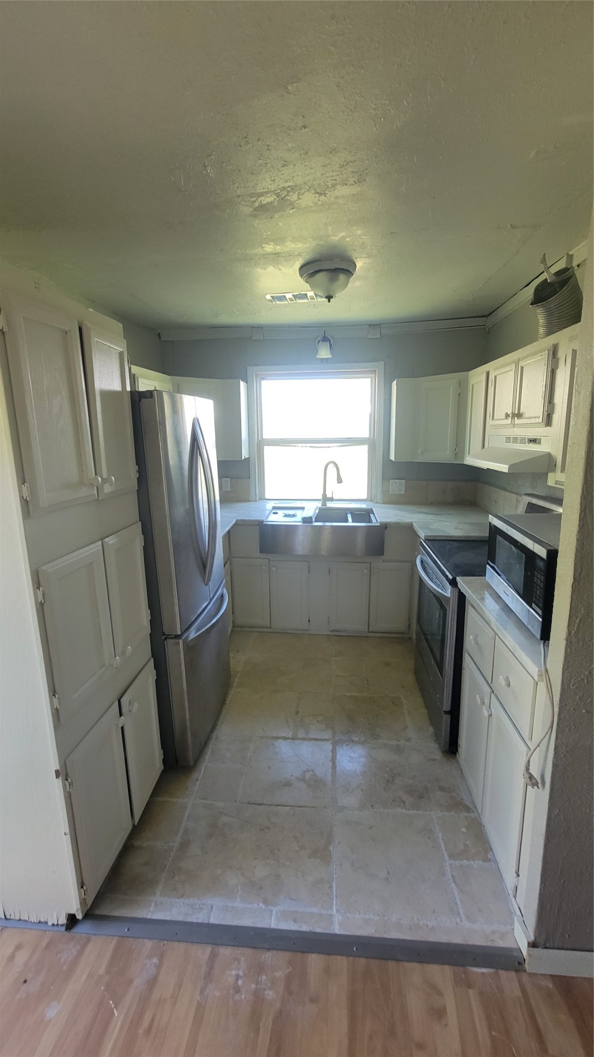 This is a compact kitchen featuring white cabinetry, a stainless steel refrigerator, and an oven. It has tiled flooring and a window above the sink for natural light, providing a cozy and functional space.