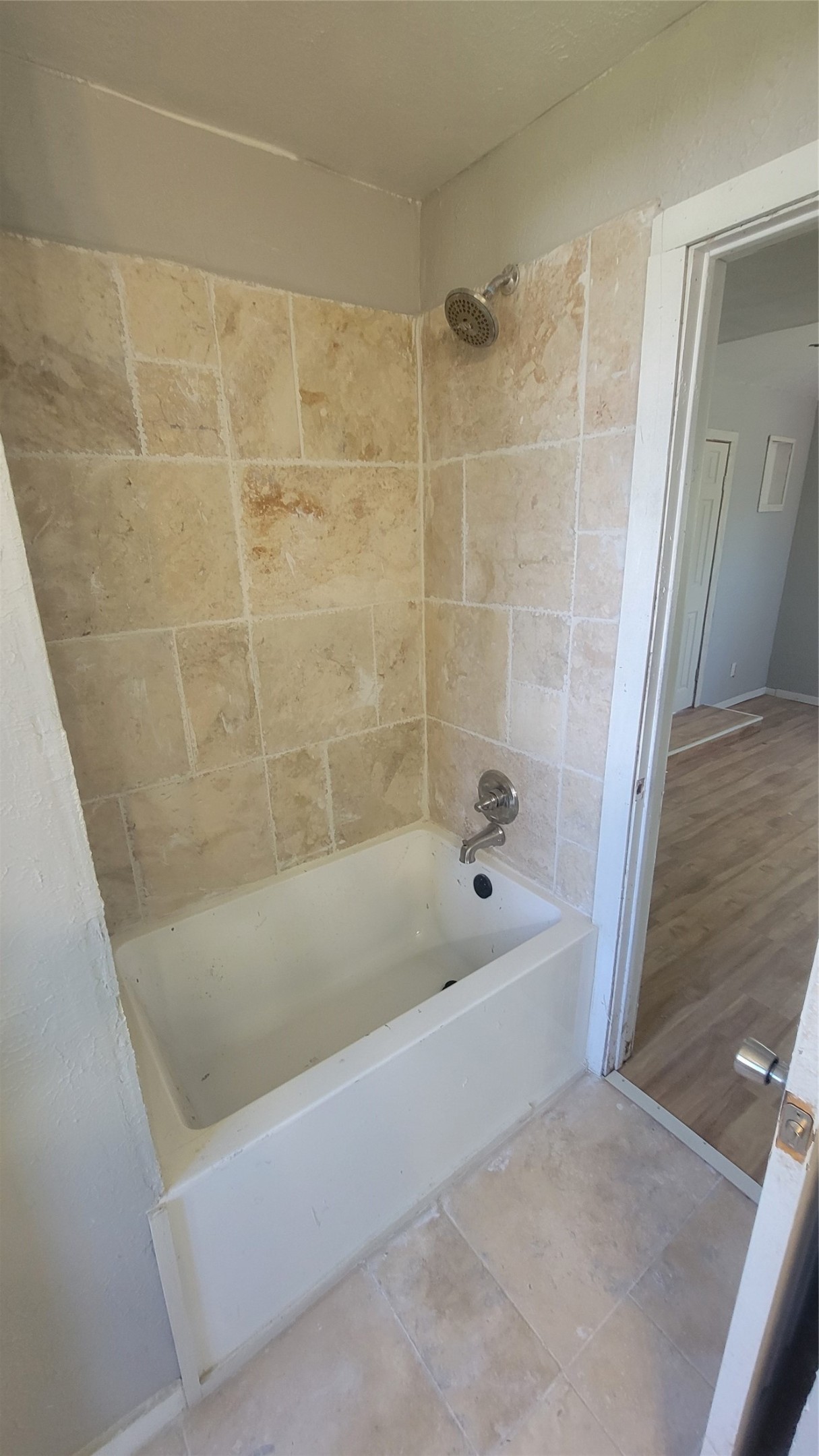 This photo shows a bathroom with a tiled shower and bathtub combination. The walls are lined with beige tiles, and the floor has matching tiles. A single showerhead is installed above the tub. The entrance to the bathroom opens to a room with wood-style flooring.