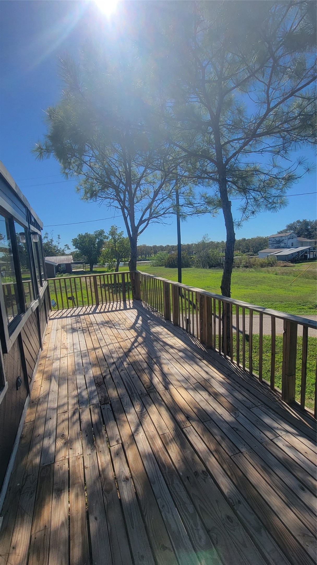 This photo showcases a spacious wooden deck with railing, offering a sunny spot for relaxation. Surrounded by greenery and trees, it provides a serene outdoor space with a view of open land and clear skies.