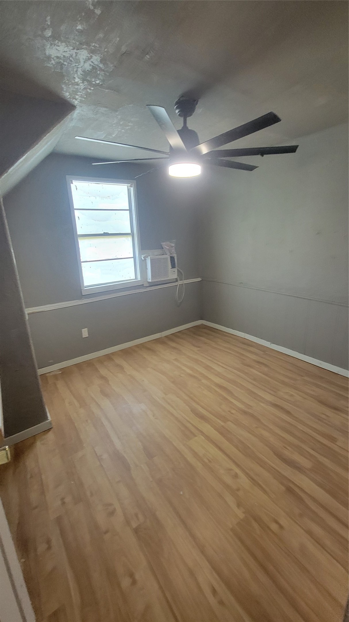 This room features laminate wood flooring, a ceiling fan with light, and a window with an air conditioning unit. It has neutral gray walls and a sloped ceiling, providing a cozy atmosphere.