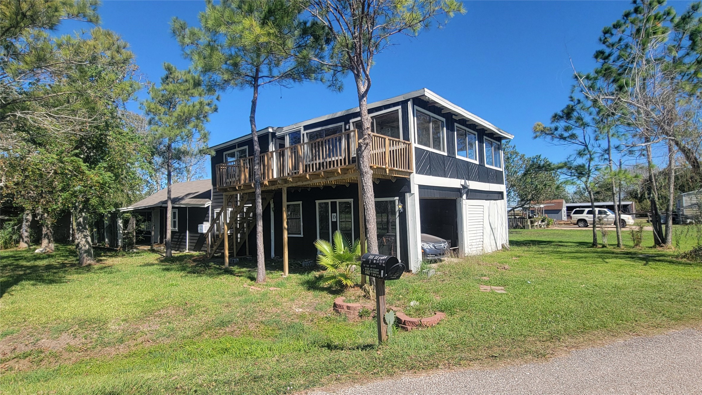 Two-story home with a wooden balcony and large windows, set in a grassy yard with mature trees. The property includes a garage and has a driveway leading to the street.