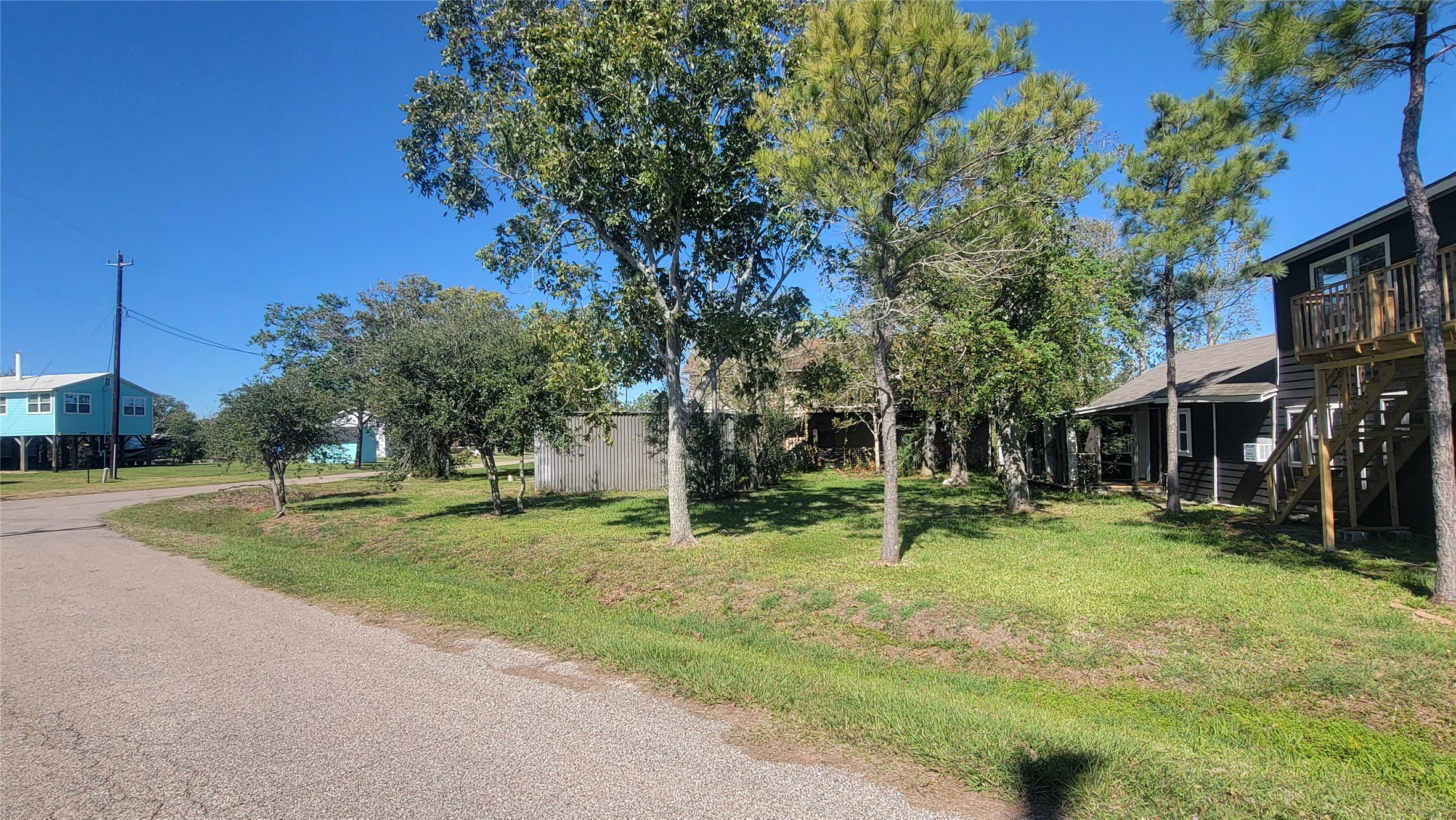 This photo shows a quiet residential street with a grassy lot and mature trees. Nearby, there are raised homes with wooden decks, suggesting a coastal or flood-prone area. The setting appears peaceful and spacious.