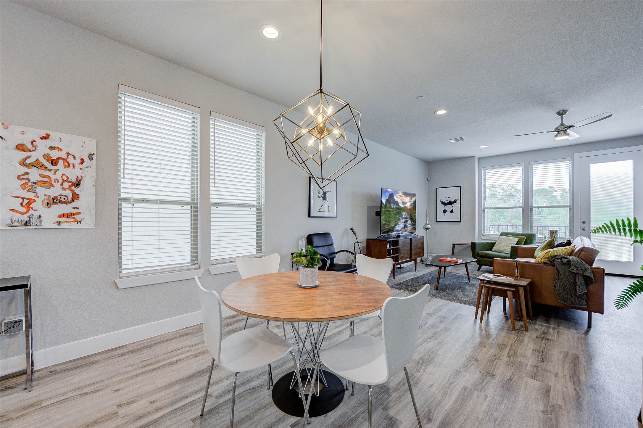 This photo showcases a modern, open-concept living and dining area with light wood flooring and ample natural light from large windows. The space features contemporary furniture, including a round dining table, stylish chairs, and a cozy seating area with a sofa and TV. A unique geometric light fixture adds a touch of elegance.