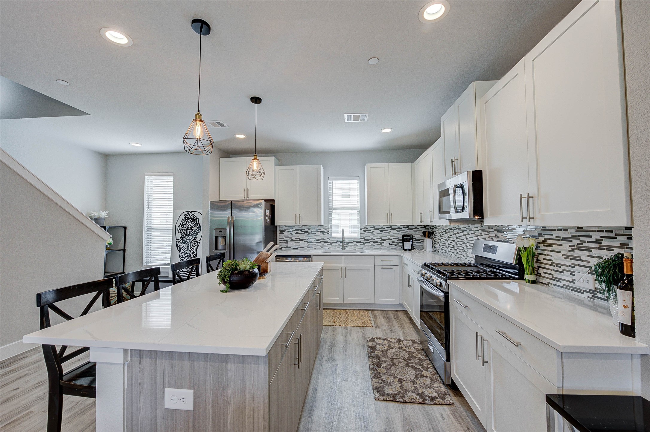 This modern kitchen features sleek white cabinetry, stainless steel appliances, and a stylish tile backsplash. The spacious island offers additional seating and workspace, while pendant lighting adds a warm touch. Natural light fills the room, highlighting the open layout and contemporary design.