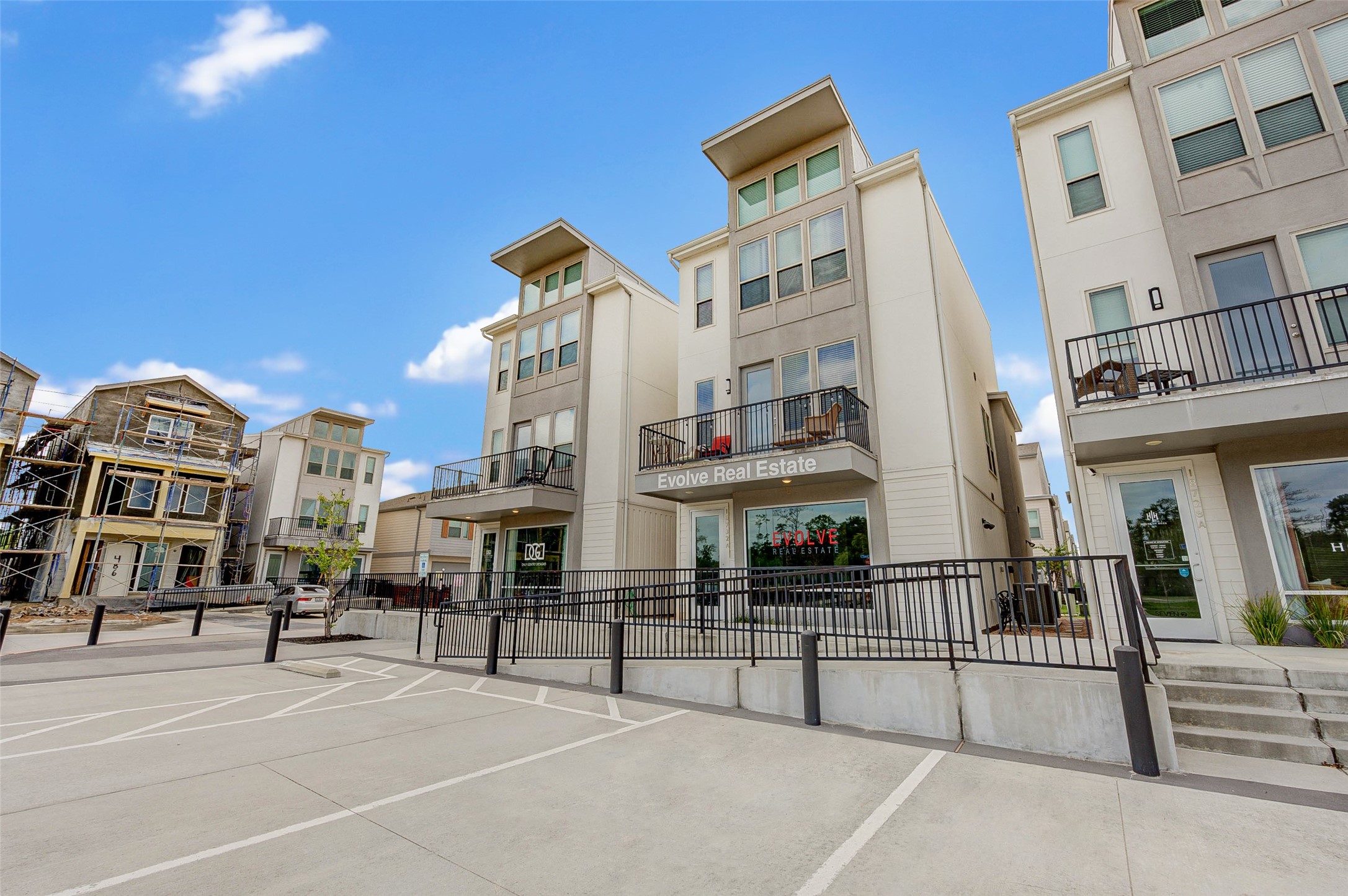 Modern multi-story townhouses with clean lines and large windows. The area features balconies, ample parking, and a clear blue sky, creating a welcoming and contemporary environment.