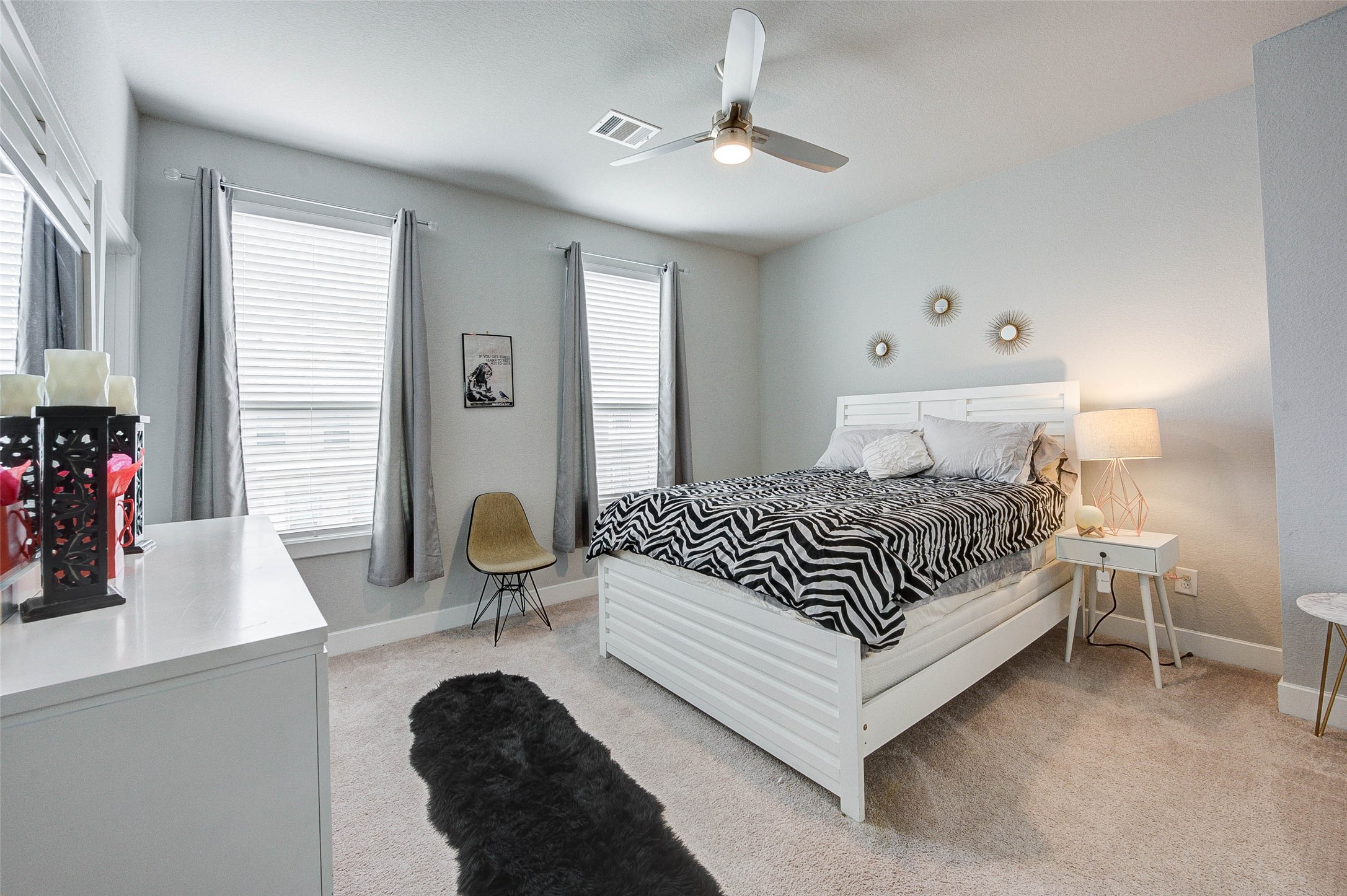 This bedroom features a cozy, modern design with soft carpet flooring and neutral walls. It includes a stylish bed with a zebra-patterned duvet, a ceiling fan, and ample natural light from two large windows with gray curtains. A chic dresser and nightstand, complemented by decorative wall accents, complete the room.