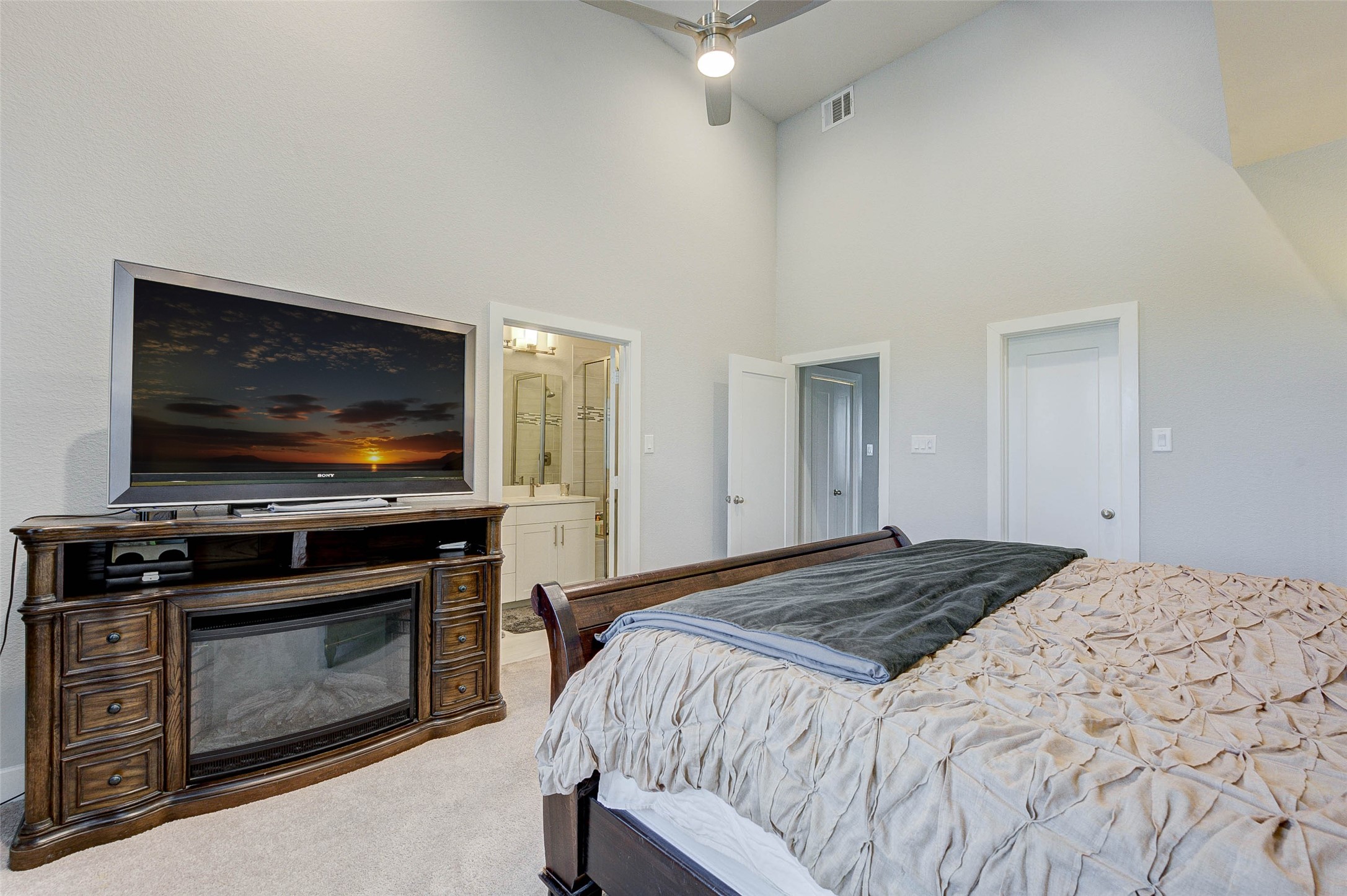 This bedroom features a cozy setup with a stylish wooden TV stand and mounted TV. It has plush carpeting, neutral walls, and a ceiling fan. There's an attached bathroom with modern fixtures, adding convenience and comfort.