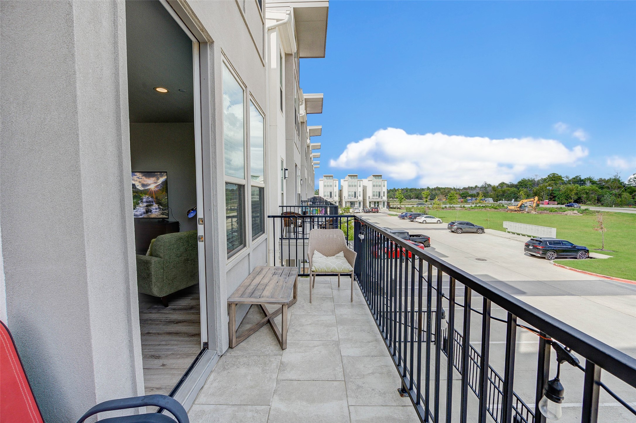 A modern balcony view featuring a spacious area with seating and a scenic outlook over a tidy street and green space. Large windows connect the outdoor and indoor spaces, offering natural light and a contemporary vibe.
