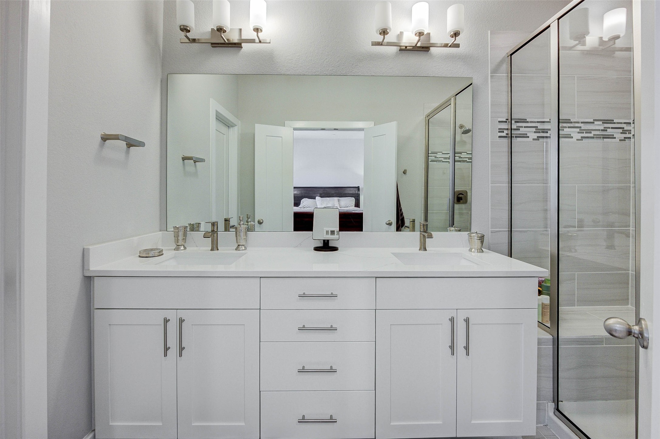 This modern bathroom features a spacious double vanity with sleek white cabinetry and dual sinks. It includes a large mirror and elegant lighting fixtures, with a glass-enclosed shower showcasing stylish tile work.