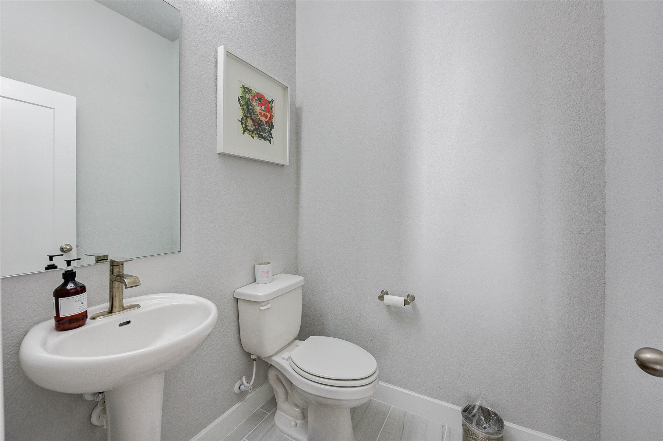 This is a modern, compact half-bathroom featuring a pedestal sink, a toilet, and neutral-toned walls. It includes a large mirror, a small wall art piece, and has a clean, minimalistic design.