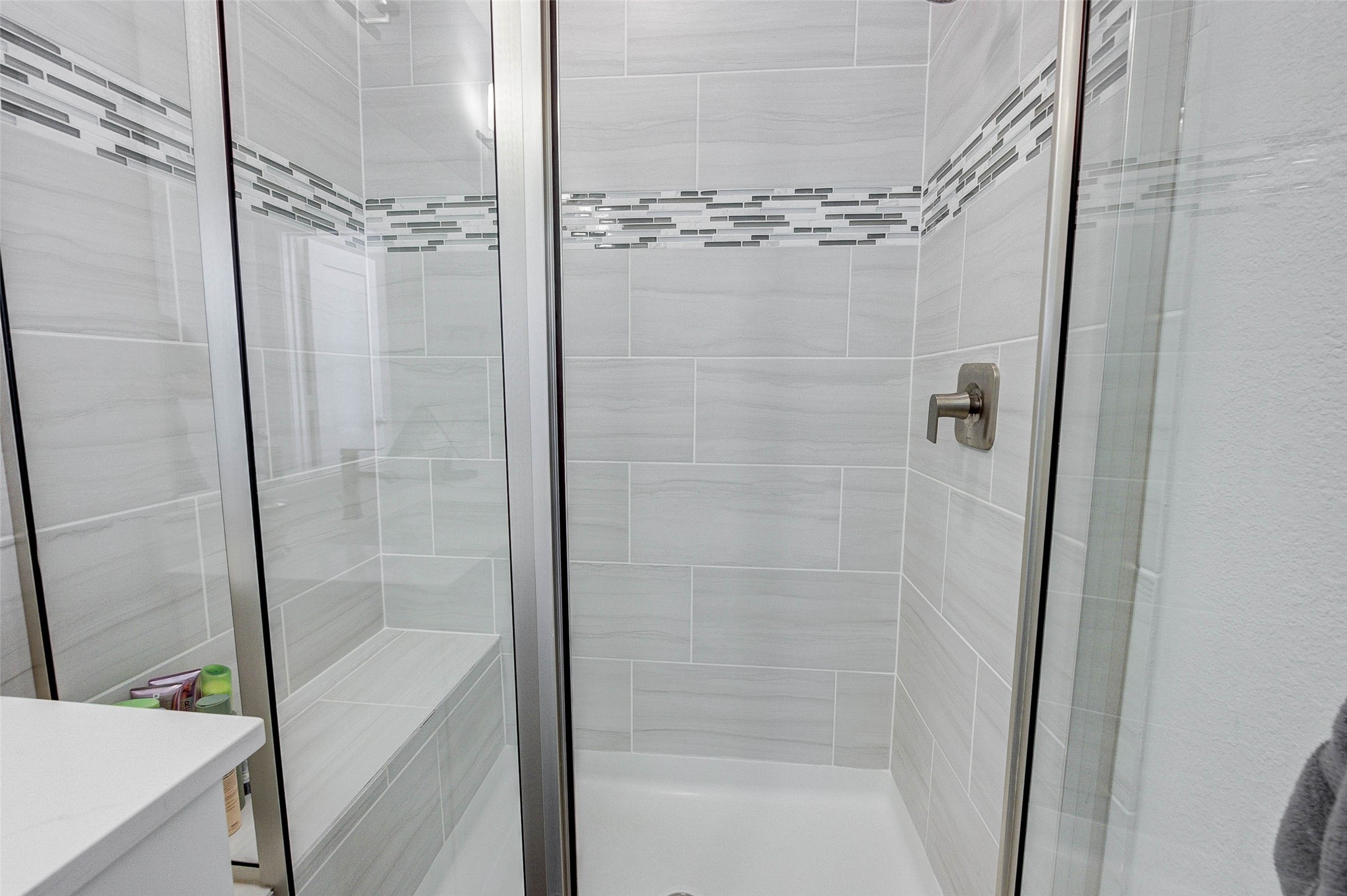 This photo shows a modern shower with sleek glass doors and a built-in bench. The walls are lined with light gray tiles accented by a decorative band of smaller, darker tiles, creating a stylish and contemporary look.