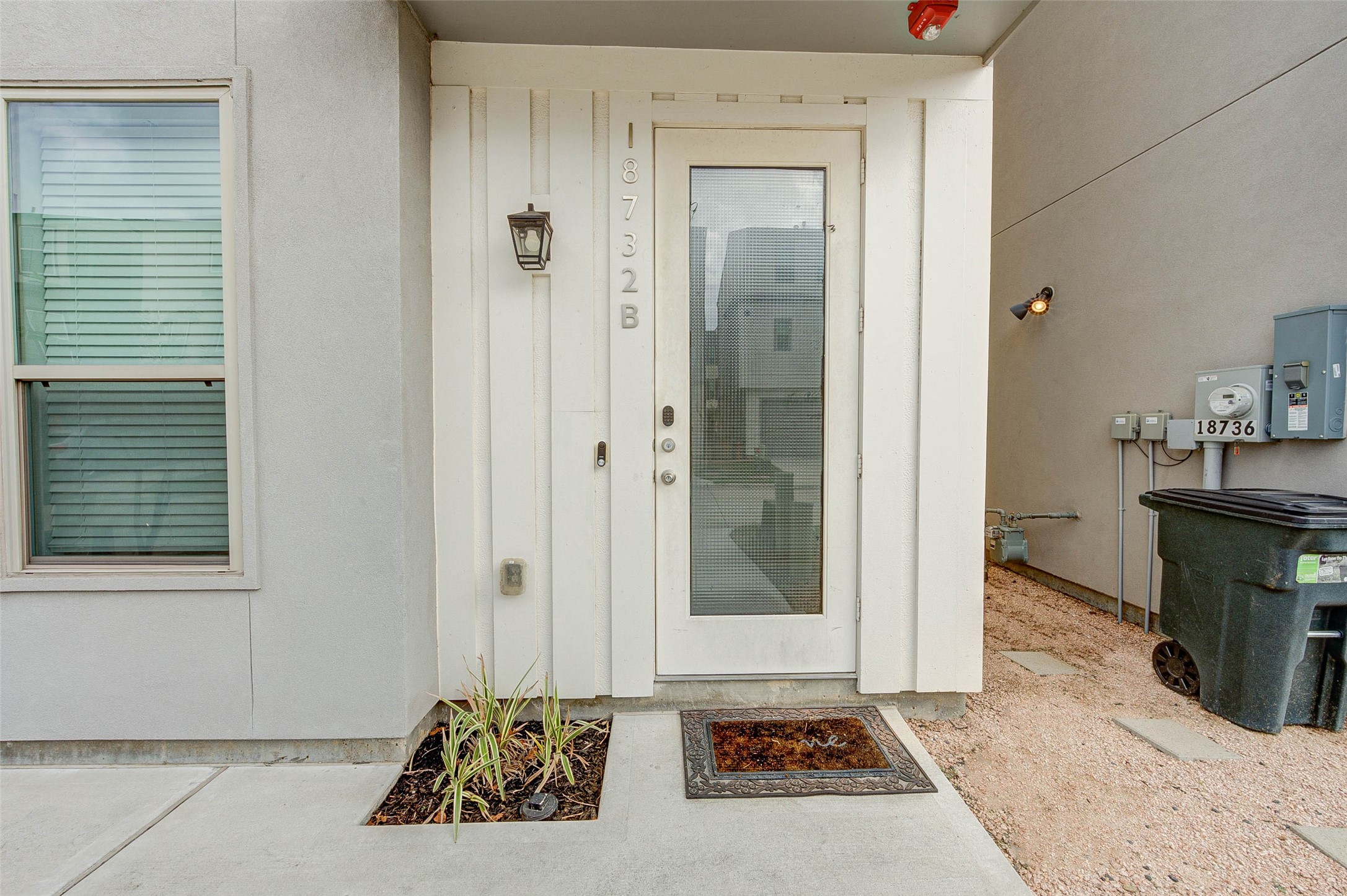 This photo shows a modern entryway with a glass-panel door and a simple exterior light fixture. The area features a small planted bed and a concrete pathway, providing a clean and welcoming entrance.
