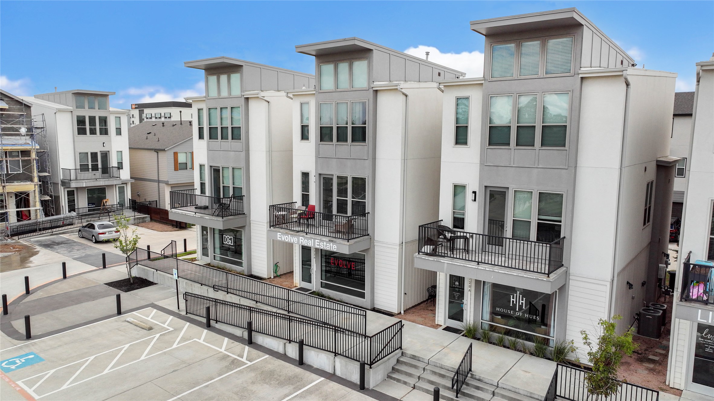 Modern townhouses with clean, contemporary architecture featuring three stories, large windows, and private balconies. The complex includes ground-level retail spaces and is situated in a well-maintained urban setting.