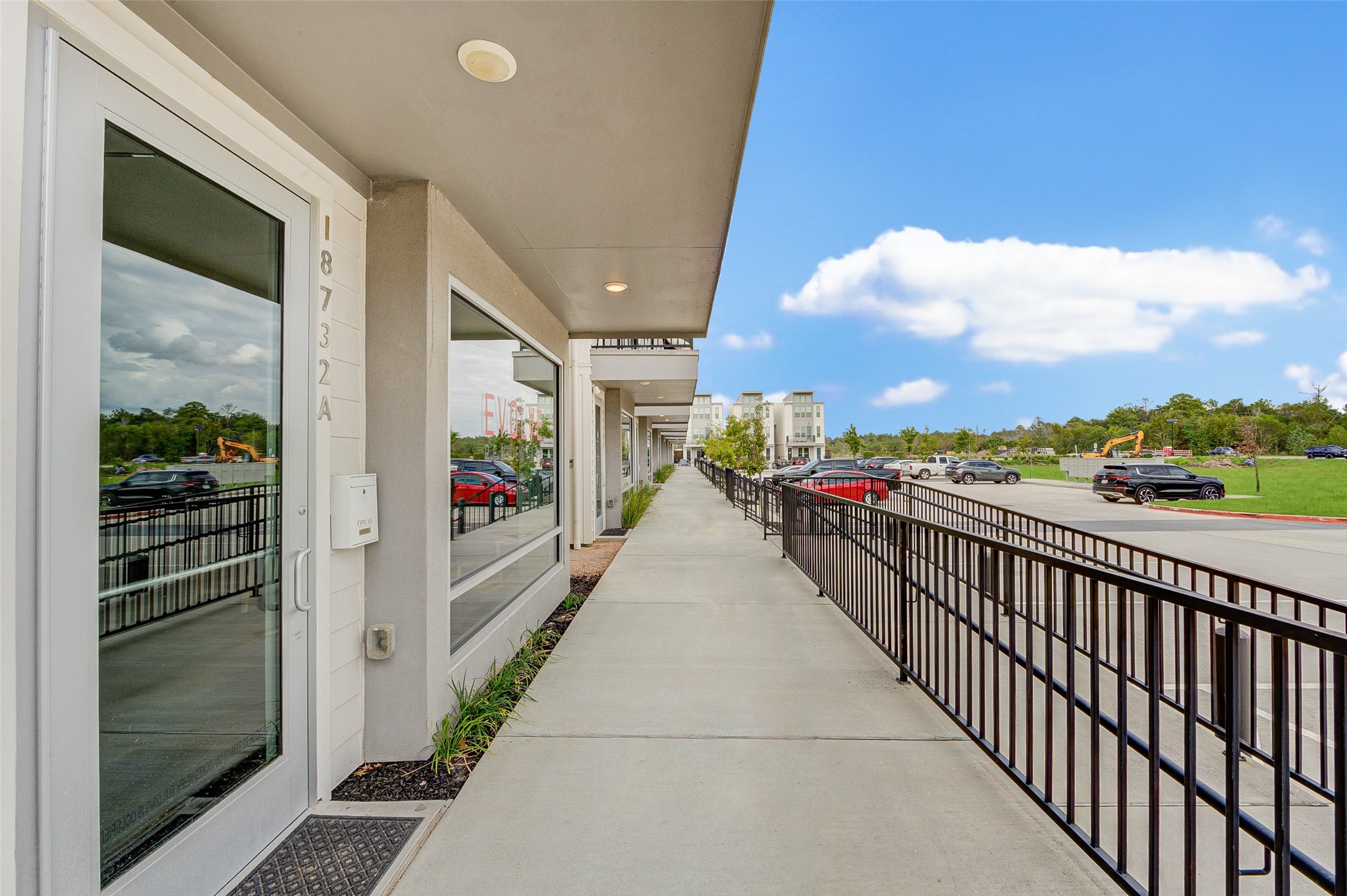 Modern building exterior with a long walkway and metal railing, adjacent to a parking lot. Glass doors and windows enhance natural light. Ideal for commercial or residential purposes in a convenient location.