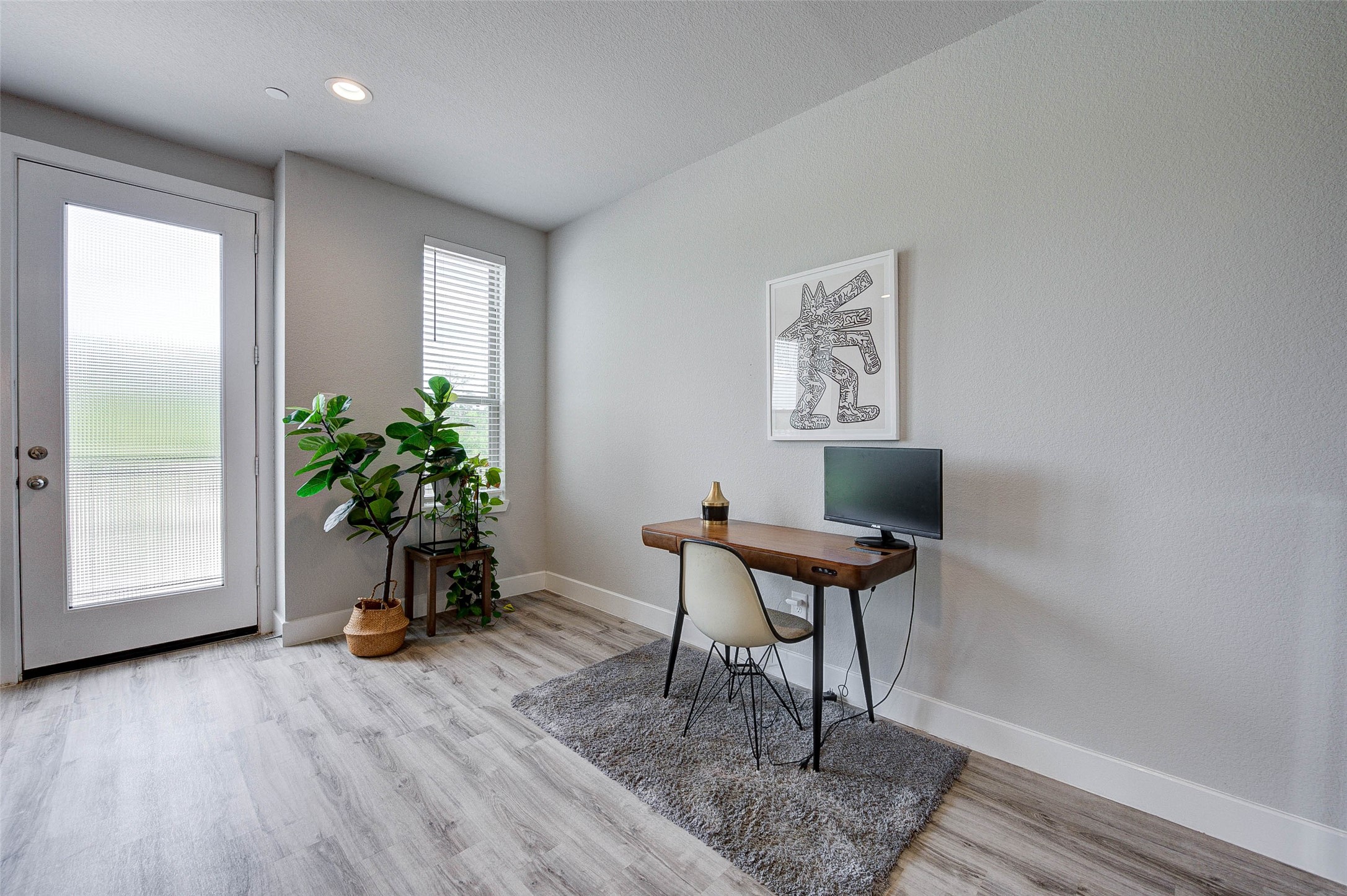 Bright and modern home office with light wood flooring, a sleek desk, and a stylish chair. Large windows and a glass door provide ample natural light, while indoor plants add a touch of greenery. Perfect for a productive work environment.