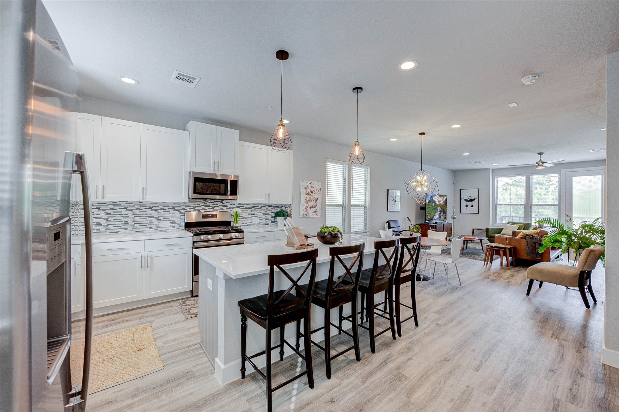Modern open-concept kitchen and living area with sleek white cabinetry, stainless steel appliances, and stylish pendant lighting. The space features a functional island with seating, light wood flooring, and ample natural light from large windows, creating a welcoming atmosphere.