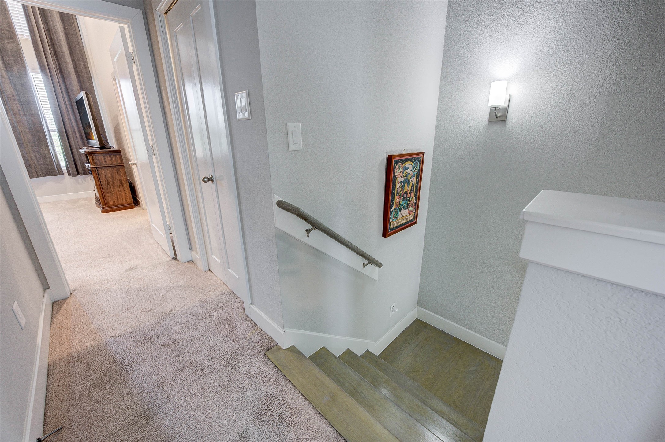 This photo shows a bright, carpeted hallway leading to a bedroom with large windows and a TV. It features a well-lit stairway with a wooden handrail and a decorative wall piece.