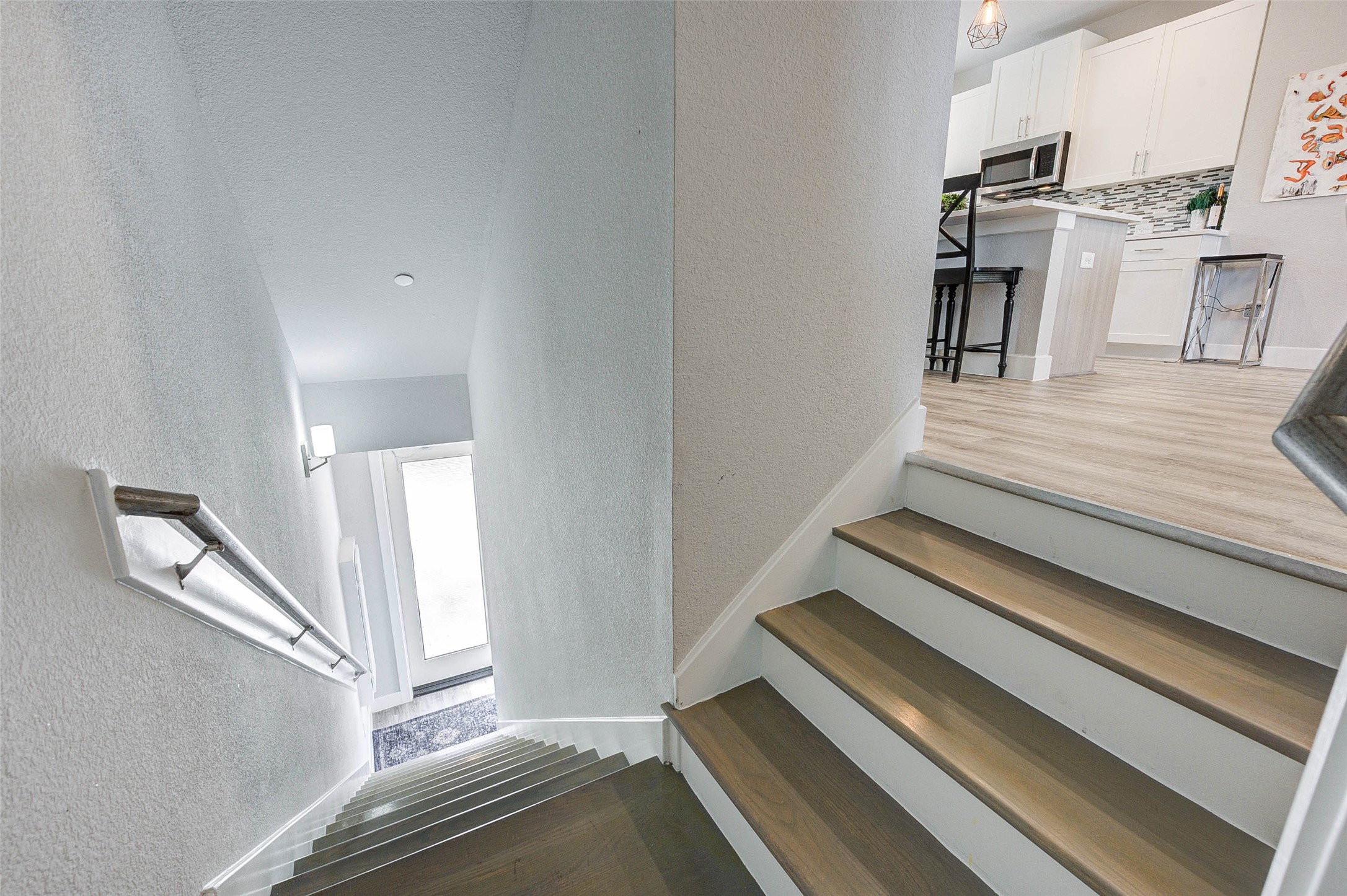 This photo shows a staircase leading to a bright, modern kitchen area with light wood flooring, white cabinetry, and a decorative backsplash. The space is open and inviting, with contemporary design elements.