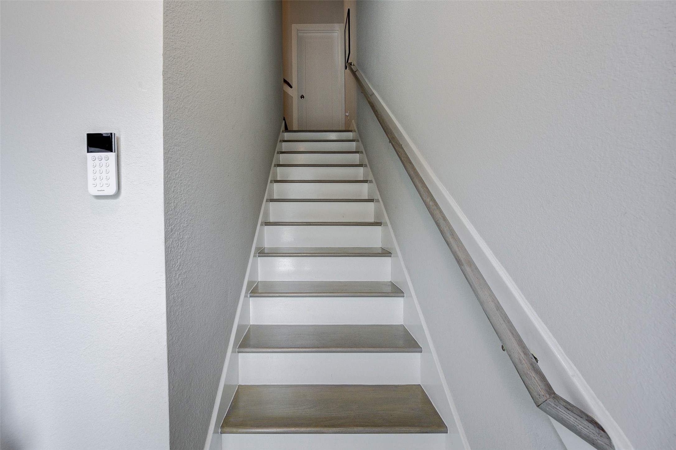 This photo shows a narrow staircase with a wooden handrail leading to a door at the top. The walls are painted white, and a security keypad is mounted on the side wall.