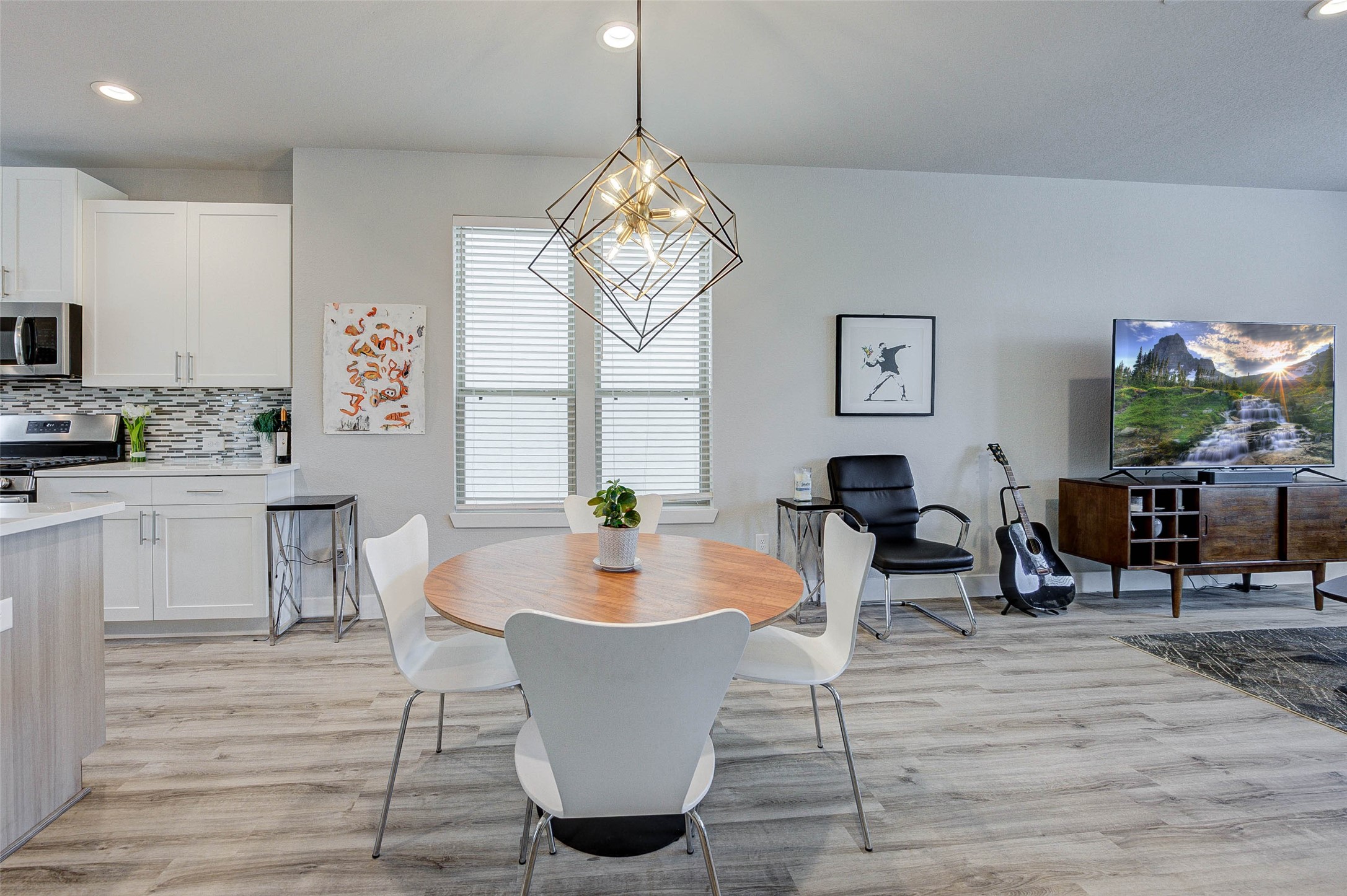 This modern open-plan living area features a sleek kitchen with stainless steel appliances and white cabinetry. It seamlessly flows into a cozy dining space with a stylish geometric light fixture. The living area includes a comfortable chair, TV setup, and contemporary decor, all set on elegant wood flooring.