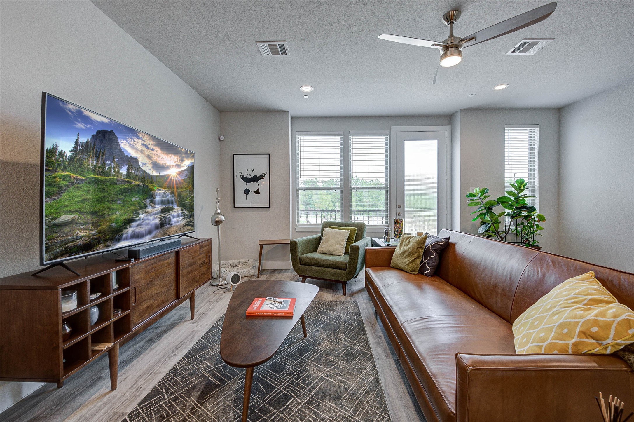 This cozy living room features a modern design with a sleek ceiling fan, a large TV, and a stylish wooden media console. The space is brightened by natural light from large windows and a door leading to an outdoor area. Comfortable seating includes a brown leather sofa and an olive armchair, complemented by a patterned rug and decorative pillows.