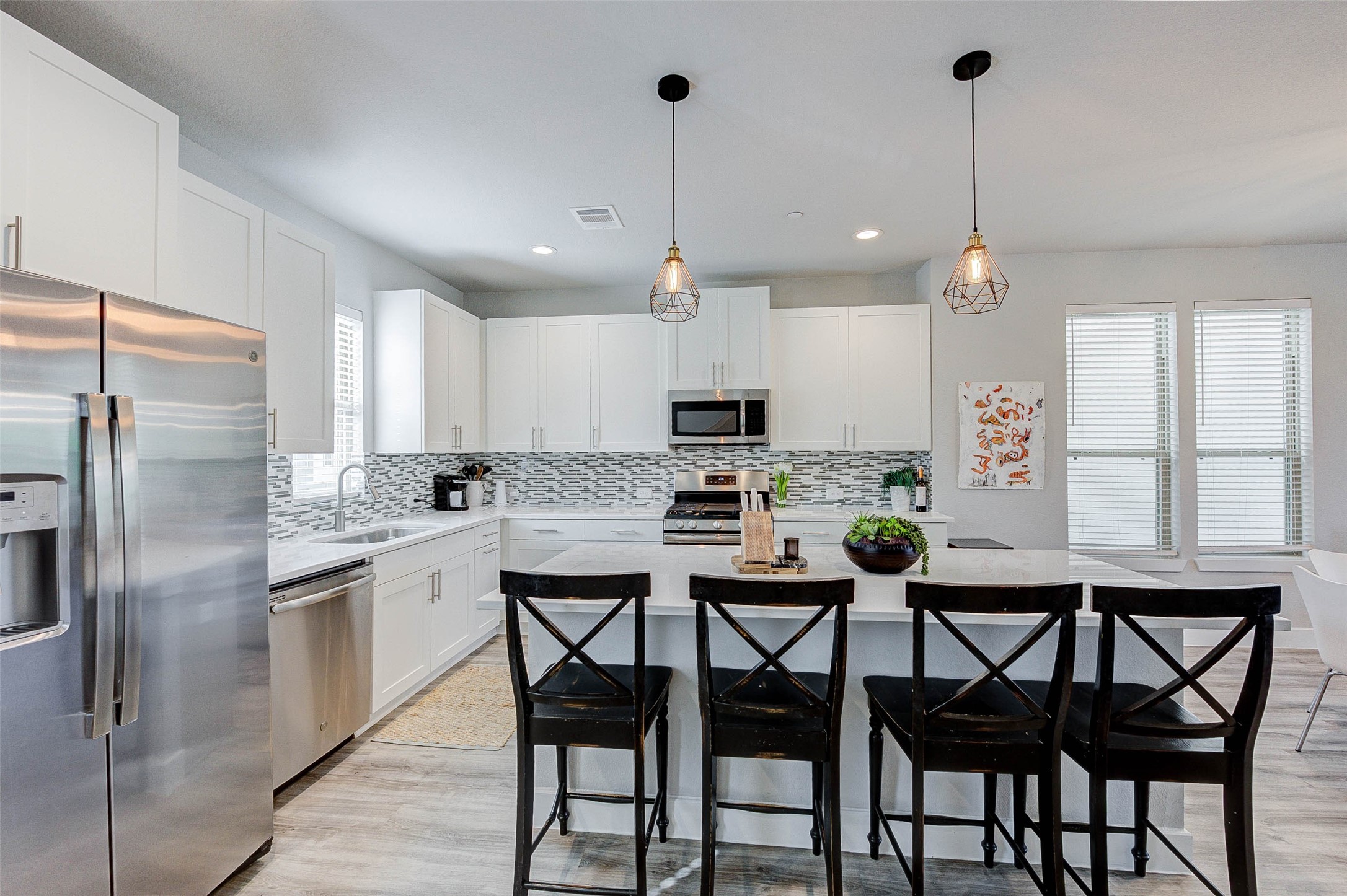 Modern kitchen with sleek white cabinetry, stainless steel appliances, and a stylish backsplash. The island features seating for four and contemporary pendant lighting. Spacious and bright with plenty of windows.