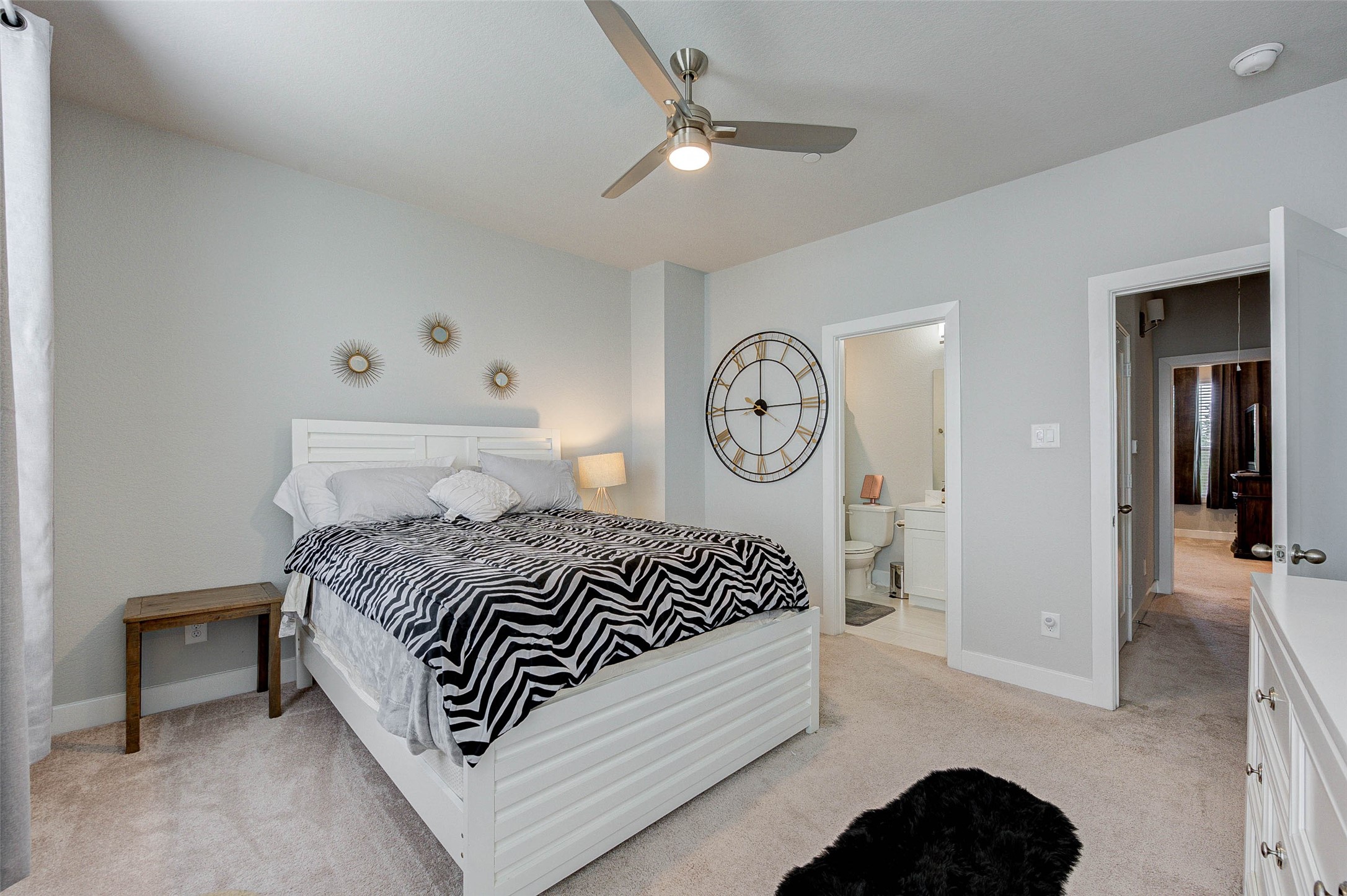 This is a cozy and bright bedroom with a modern ceiling fan, neutral walls, and carpeted flooring. It features a large wall clock, stylish decor, and an en-suite bathroom, providing both functionality and style.