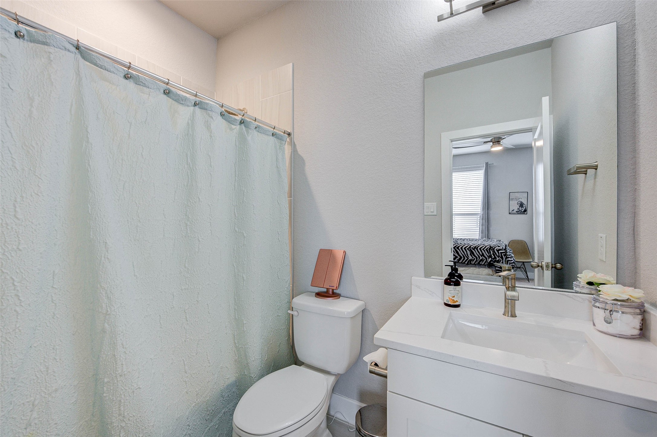 This bathroom features a clean, modern design with a white vanity and sink, complemented by a large mirror. It includes a toilet and a shower with a light-colored curtain. The room is well-lit, enhancing its fresh and airy feel.