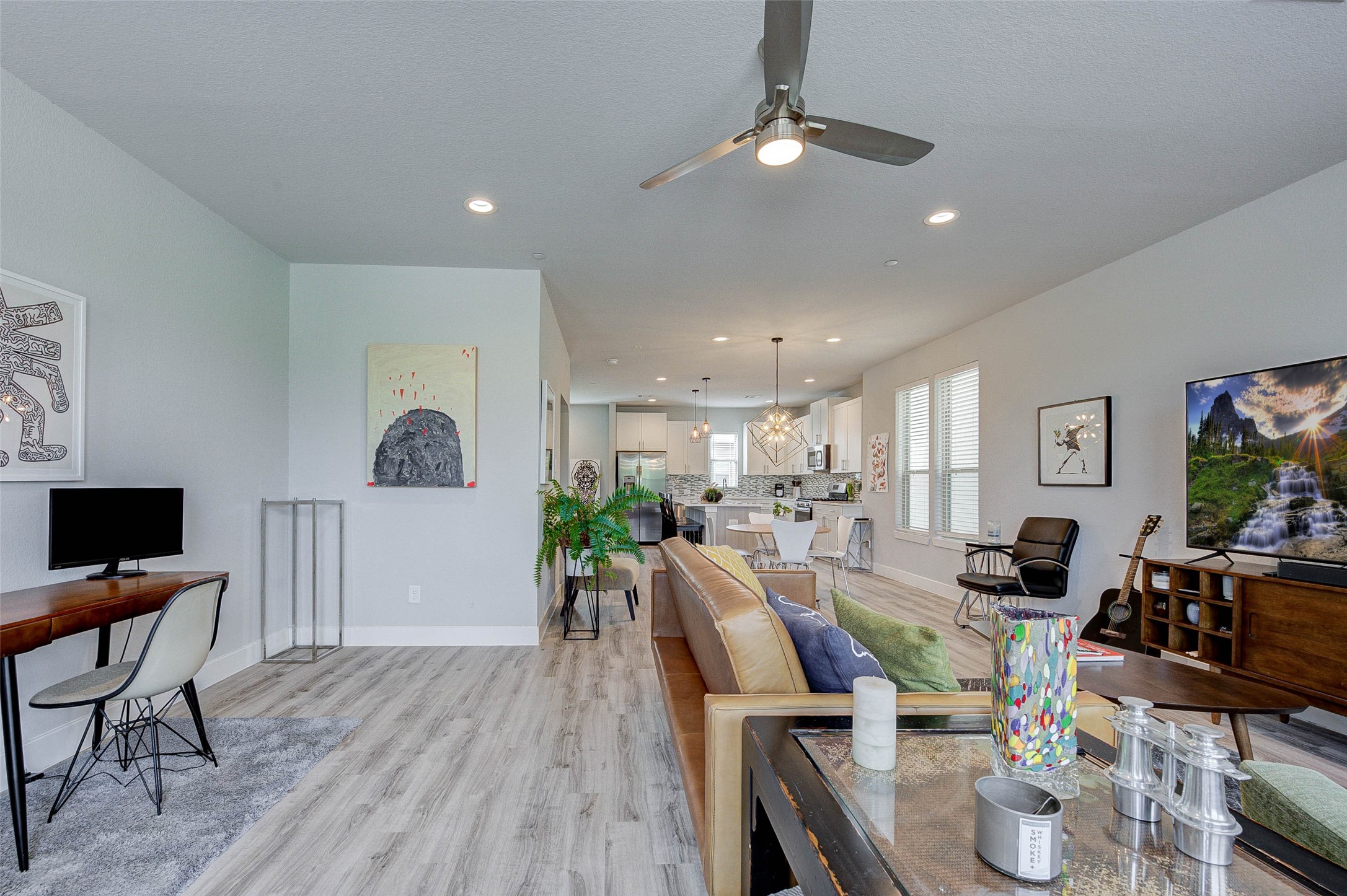 This modern open-concept living space features a cozy seating area with a stylish ceiling fan, a work desk, and a large TV. The kitchen is in the background with sleek cabinetry, stainless steel appliances, and a central island. The room is well-lit with natural light from multiple windows, and there are tasteful decorative touches throughout.