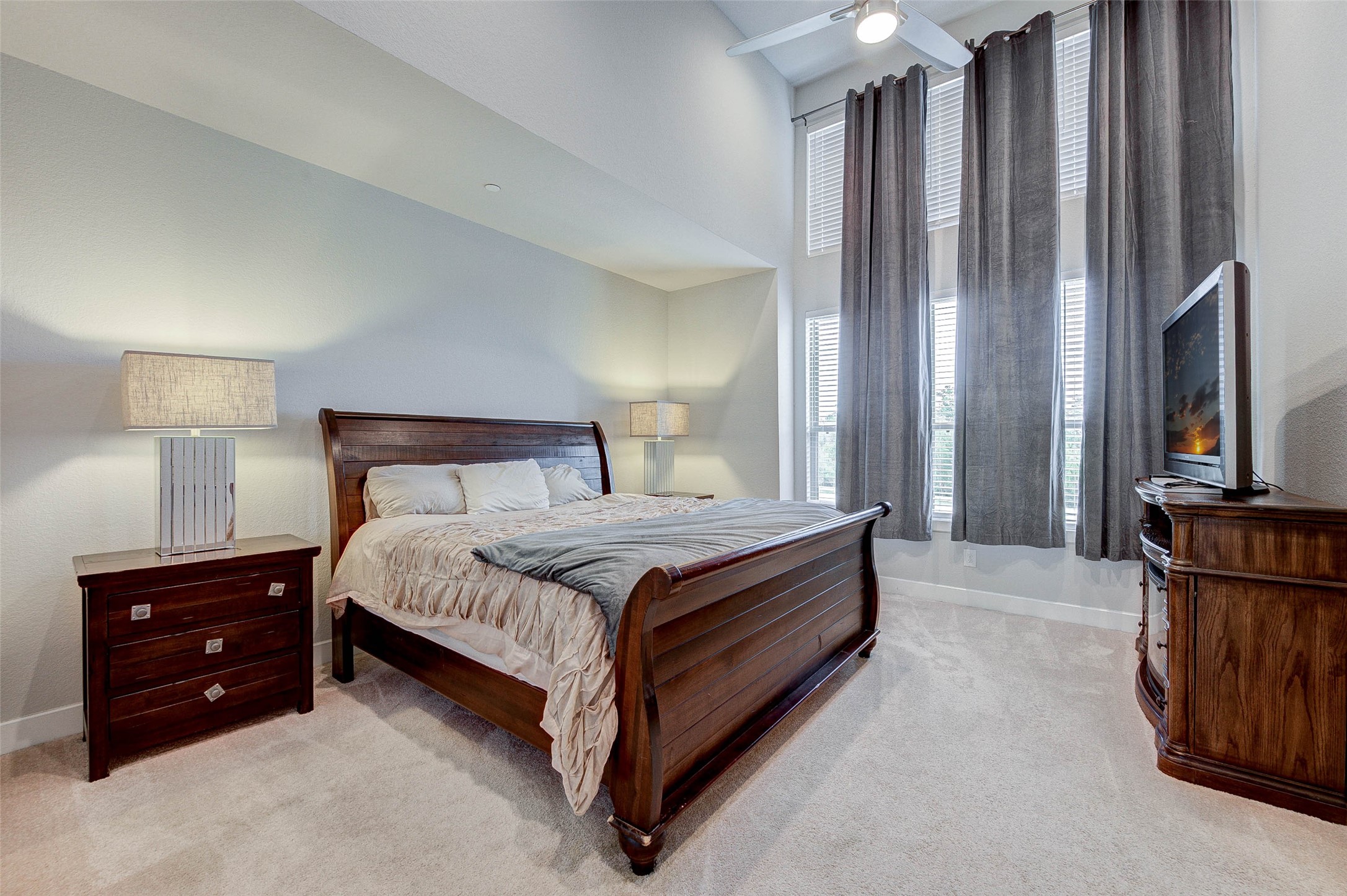 This spacious bedroom features a high ceiling, large windows with elegant drapes, and abundant natural light. It includes a wooden bed set with matching nightstands and a dresser, creating a warm and inviting atmosphere.