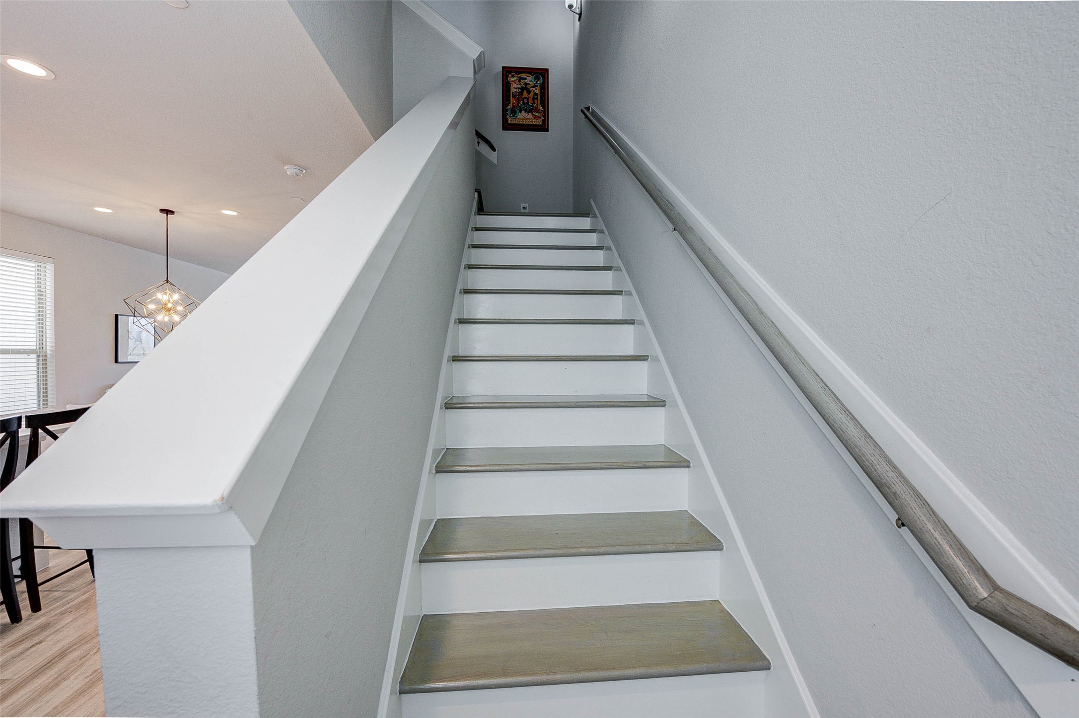 This photo shows a modern staircase with light gray steps and a sleek handrail. The walls are painted a soft gray, and there's a glimpse of a dining area with contemporary lighting. A small piece of artwork hangs at the top of the stairs.
