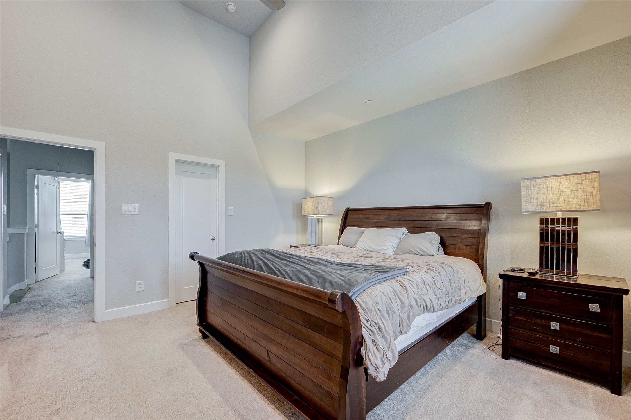 This spacious bedroom features a high ceiling and soft carpeting, creating an inviting atmosphere. It includes a large wooden bed with matching nightstands and modern lamps, offering a comfortable and stylish retreat. A doorway leads to additional living spaces, enhancing the room's functionality.
