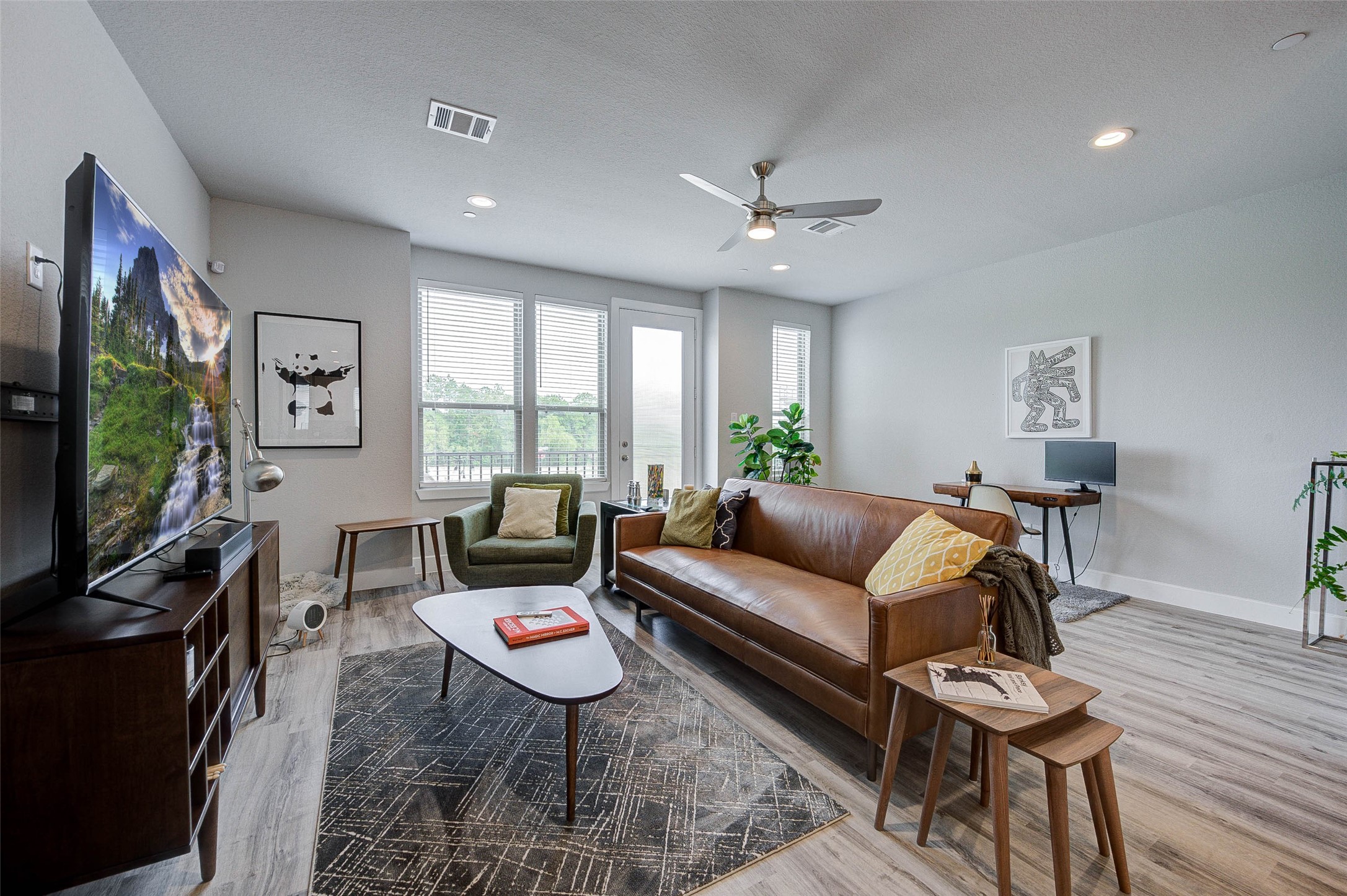 Bright and modern living room with large windows, featuring a stylish leather sofa, sleek TV setup, and a small work desk. The space is accented with plants and contemporary artwork, offering a cozy yet sophisticated atmosphere.