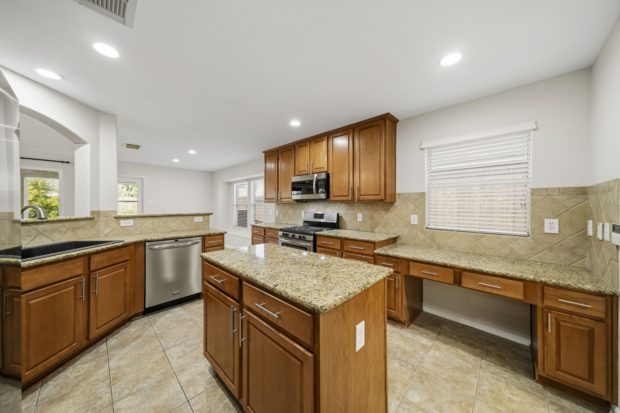 Kitchen features a desk area with a large window, tile backsplash, modern finishes, and stainless steel appliances. This bright space is perfect for work-from-home tasks, offering ample natural light and a seamless transition between culinary prep and productivity.