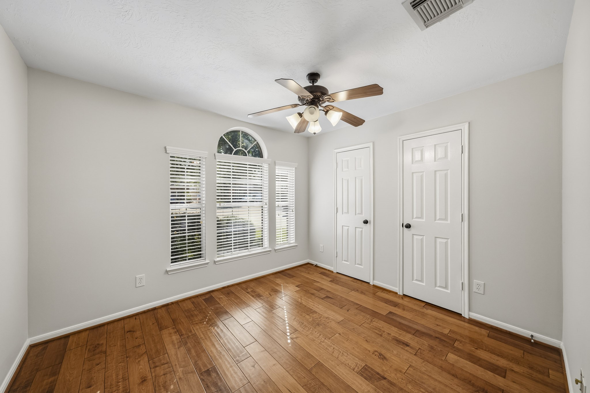 Spacious secondary bedroom featuring gleaming hardwood floors, dual closets, a modern lighted ceiling fan, and elegant windows accentuated by a sophisticated architectural arch.