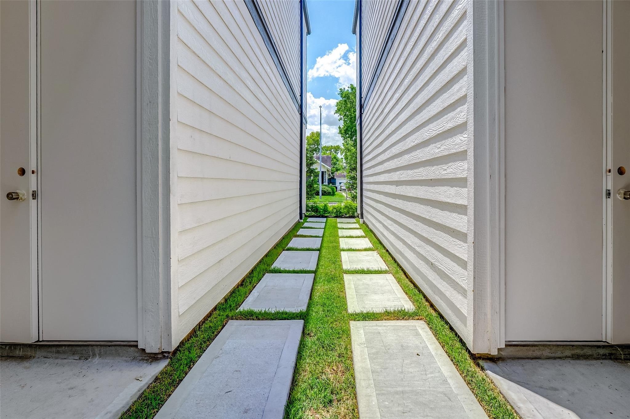 Charming walkway nestled between two buildings, featuring sleek horizontal siding and a modern path of concrete slabs set in lush green grass.