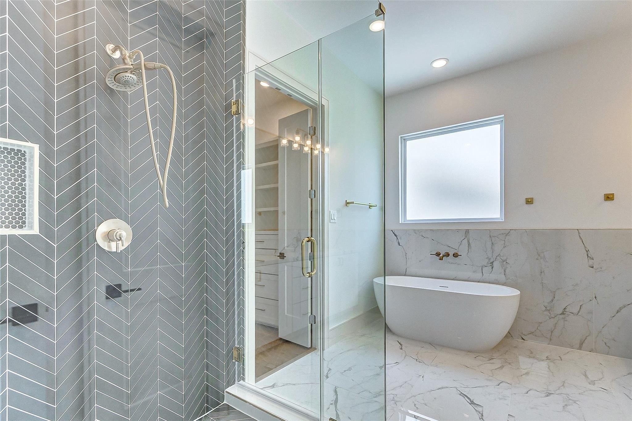 Upscale bathroom featuring a luxurious freestanding soaking tub, modern glass-enclosed frameless shower with sleek herringbone tiles, linen closet, dual vanities, large mirror, very spacious walk in closet, private commode, and bright natural light through a frosted window.