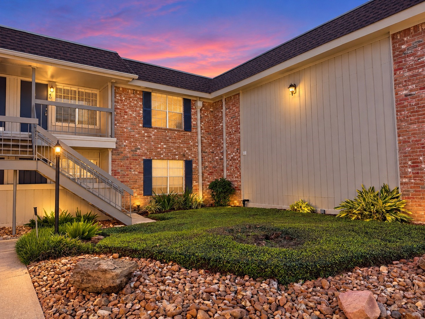 Twilight Enhanced: Charming brick exterior overlooking a landscaped courtyard with mature greenery and decorative stone accents. Twilight-enhanced photography highlights the welcoming exterior and second-floor access.