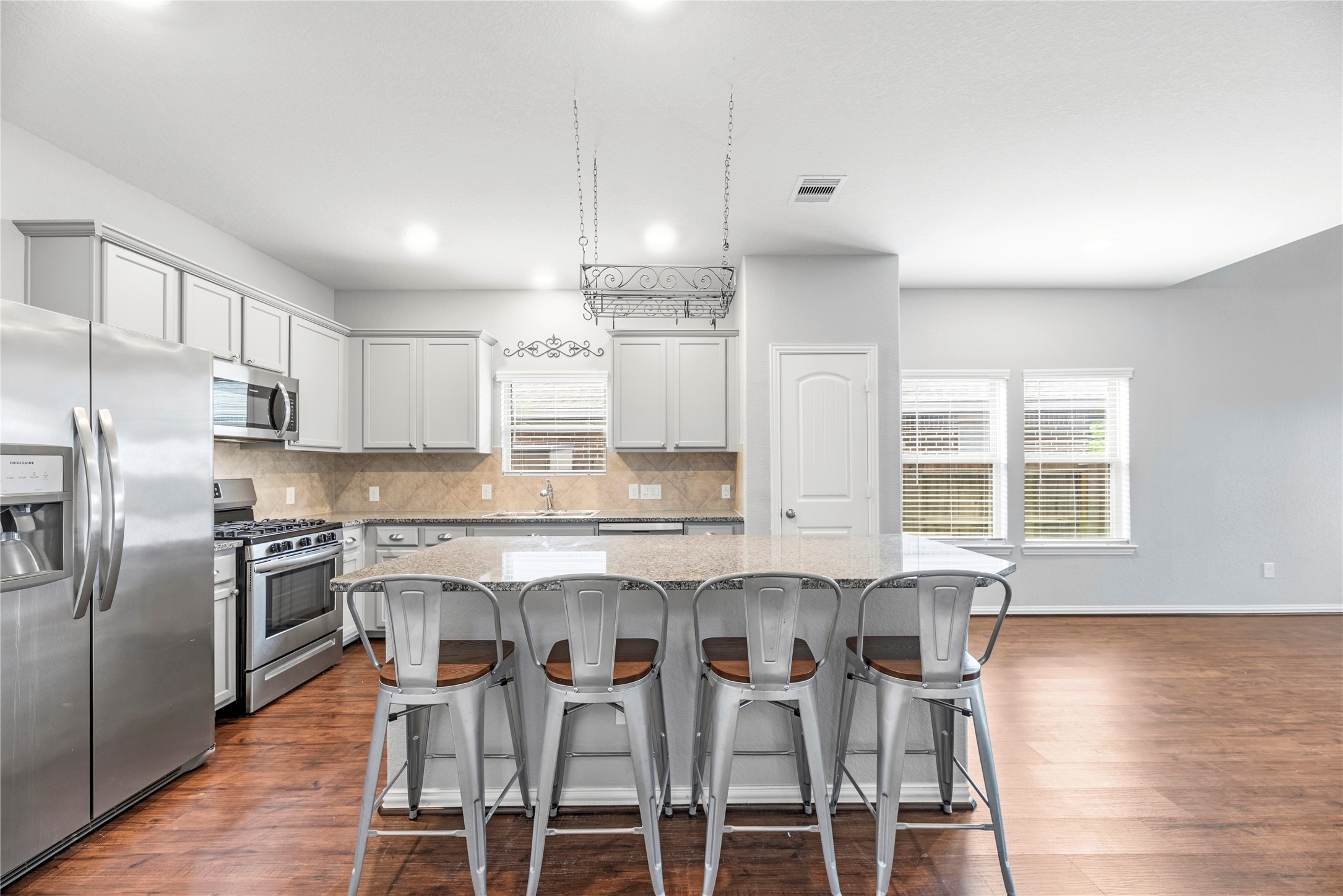 The kitchen features  granite countertops, a massive central island with breakfast bar seating, light gray cabinetry with modern hardware, and high-end stainless steel appliances, including a gas cooktop and oven.