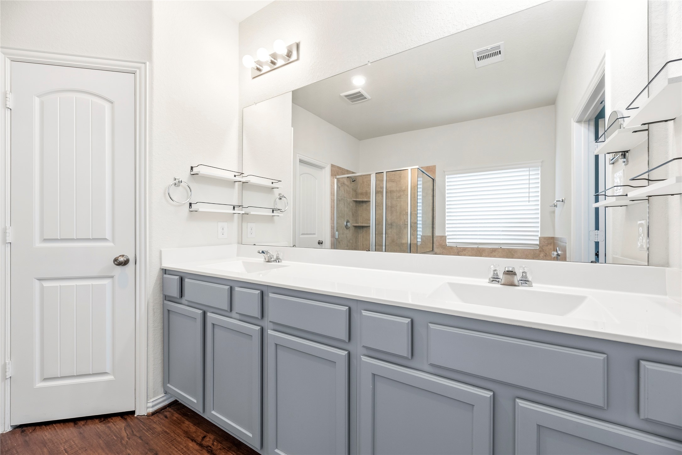 The primary bathroom showcases a extended custom vanity finished with cool gray cabinetry, dual integrated sinks, and polished hardware. A wall-to-wall mirror reflects the bright multi-bulb vanity lighting across the space.