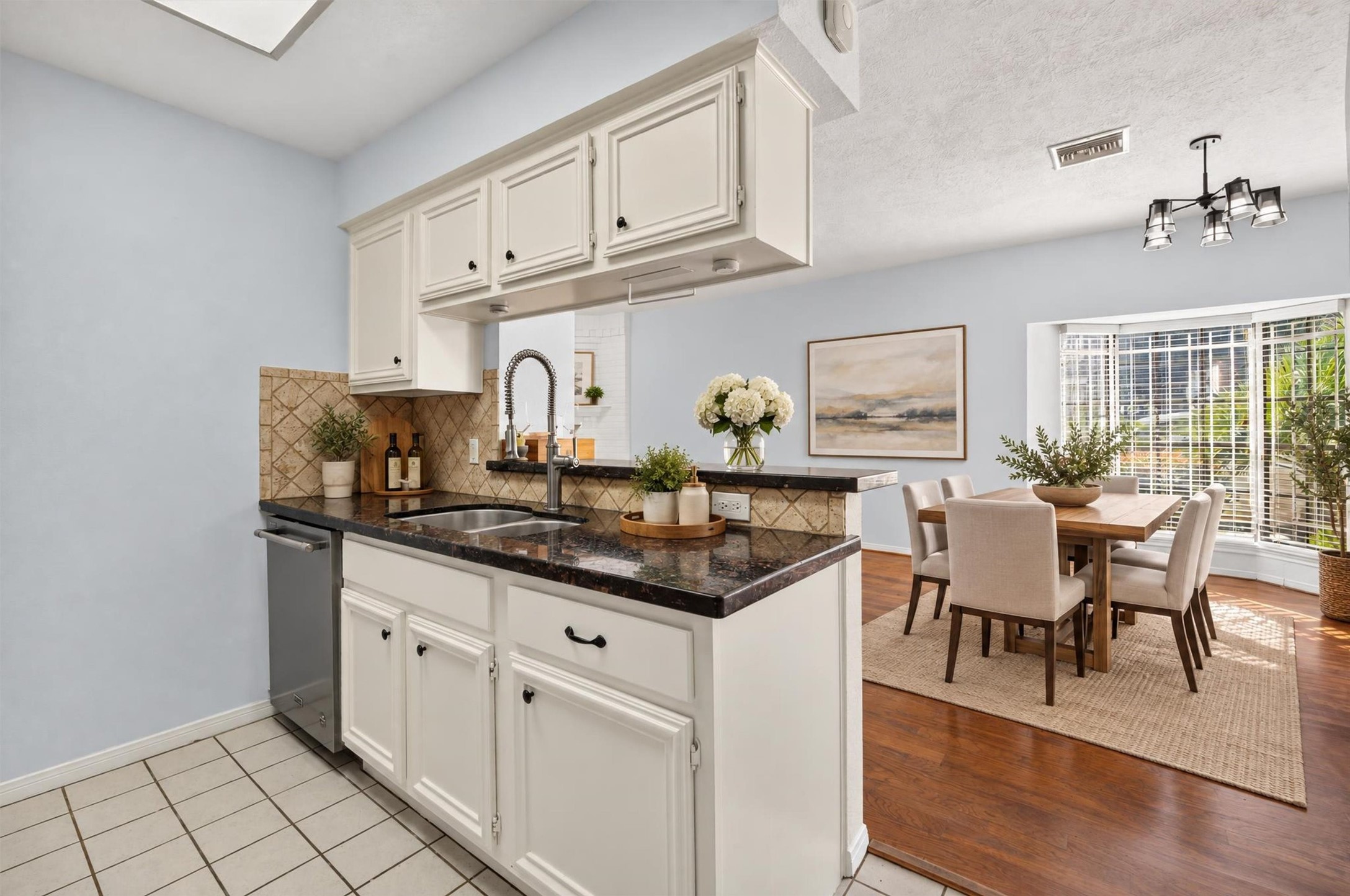 Bright kitchen with white cabinetry, granite countertops, a stainless steel dishwasher, and a breakfast bar opening to a sunlit dining area with a bay window. Virtually staged.