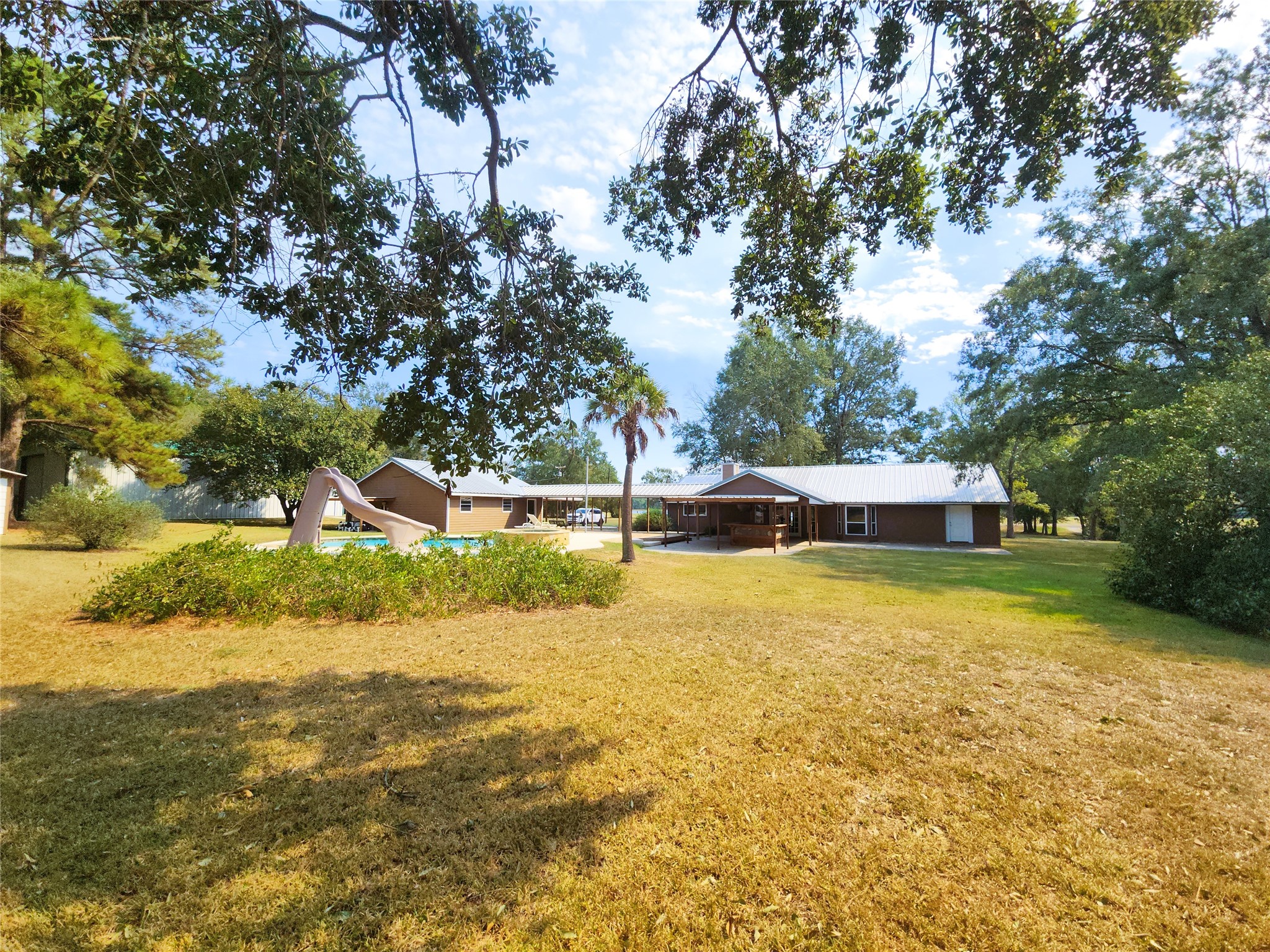 Expansive backyard featuring a charming home with a metal roof, surrounded by mature trees and a private pool with a slide, perfect for outdoor enjoyment.