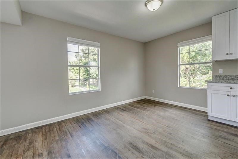 Inviting room with large windows, sleek hardwood flooring, and a glimpse of a modern kitchen area featuring white cabinetry.